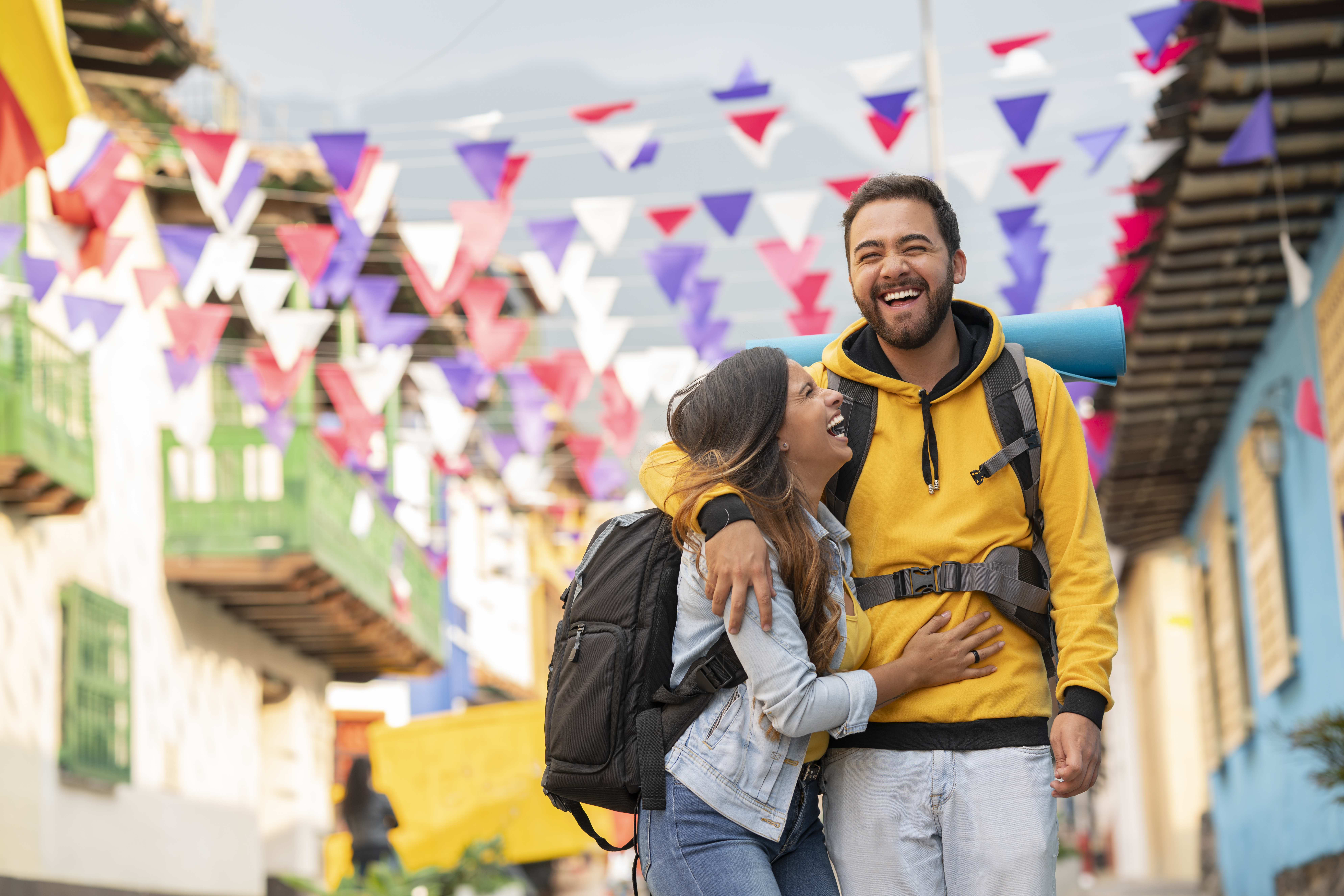 A joyful couple with backpacks laugh together in a colorful street decorated with festive flags.