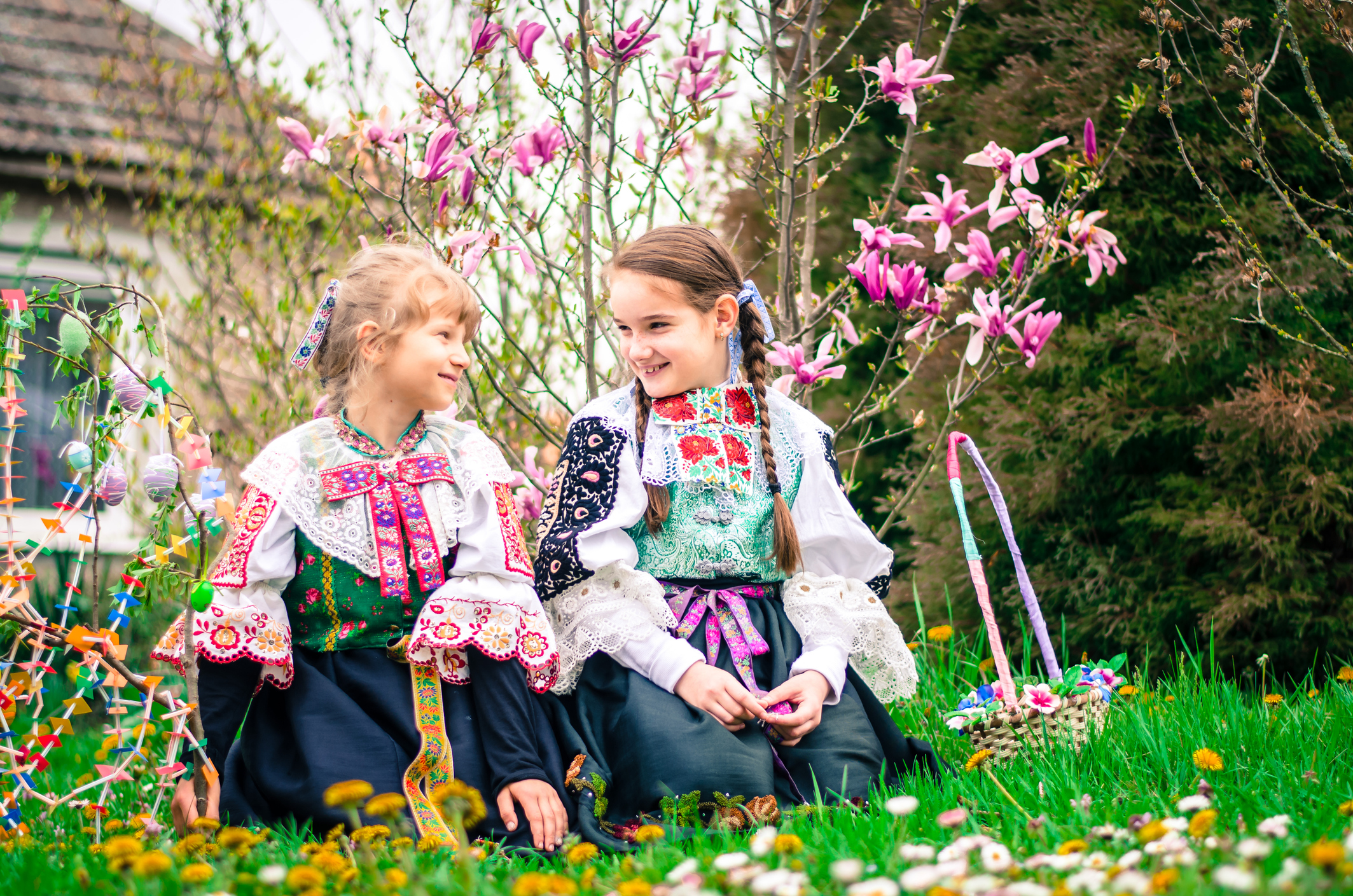 Two girls in traditional costumes sit on the grass, smiling among blooming flowers and plants.