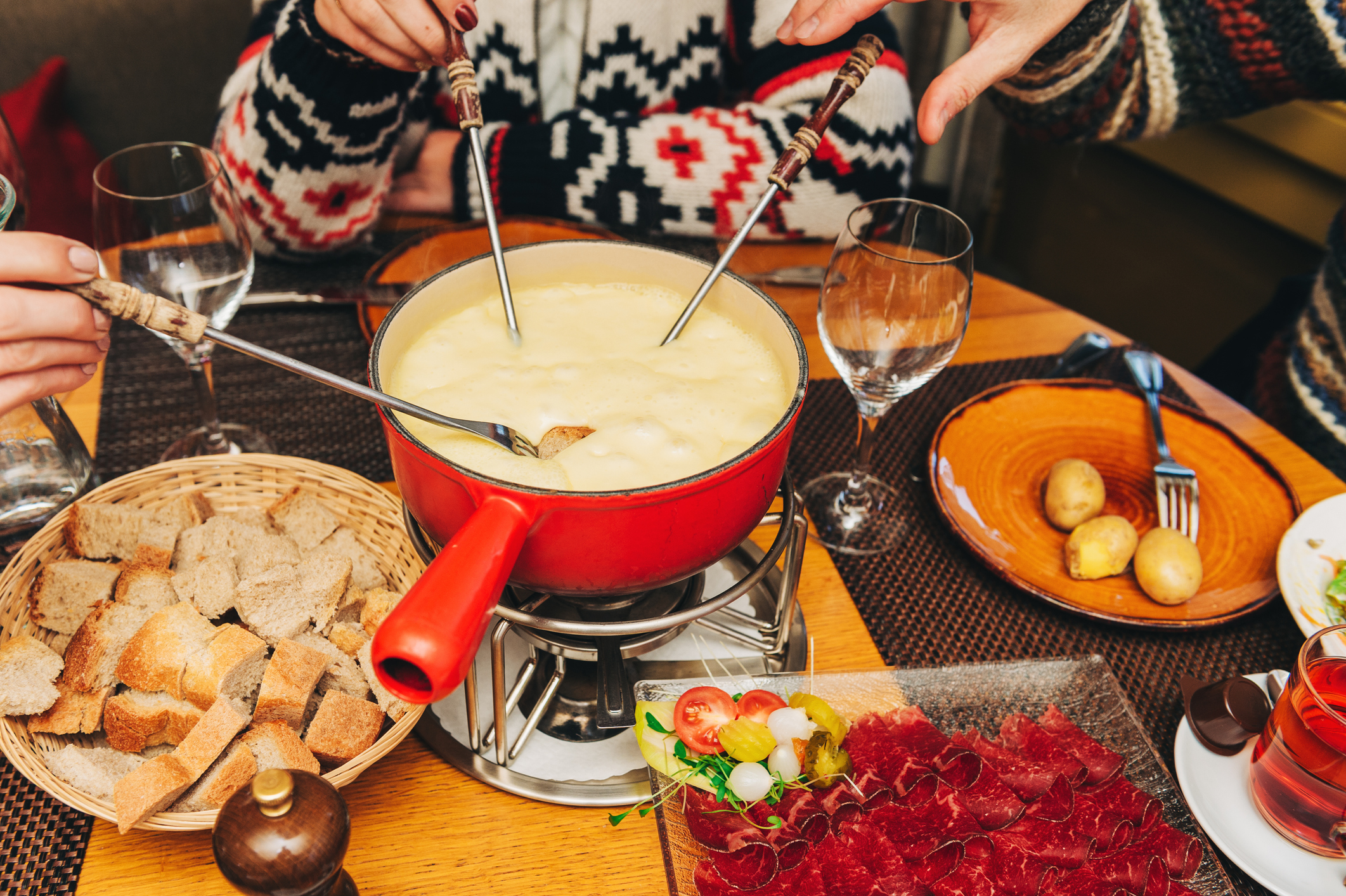 A cheese fondue pot surrounded by bread, vegetables, and drinks, with hands dipping into the fondue.
