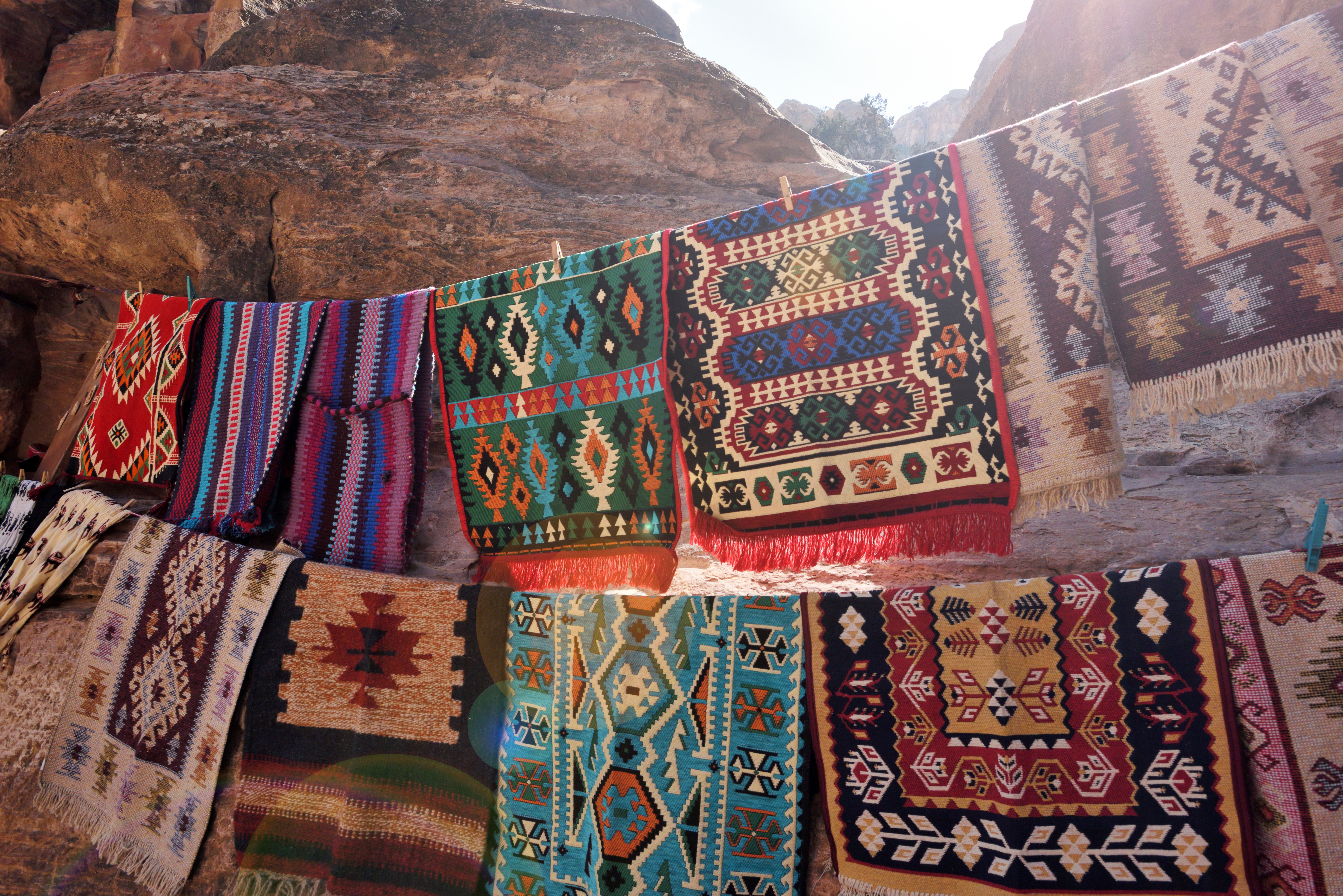 Colorful woven rugs displayed on a rocky surface, showcasing intricate patterns and textures in sunlight.