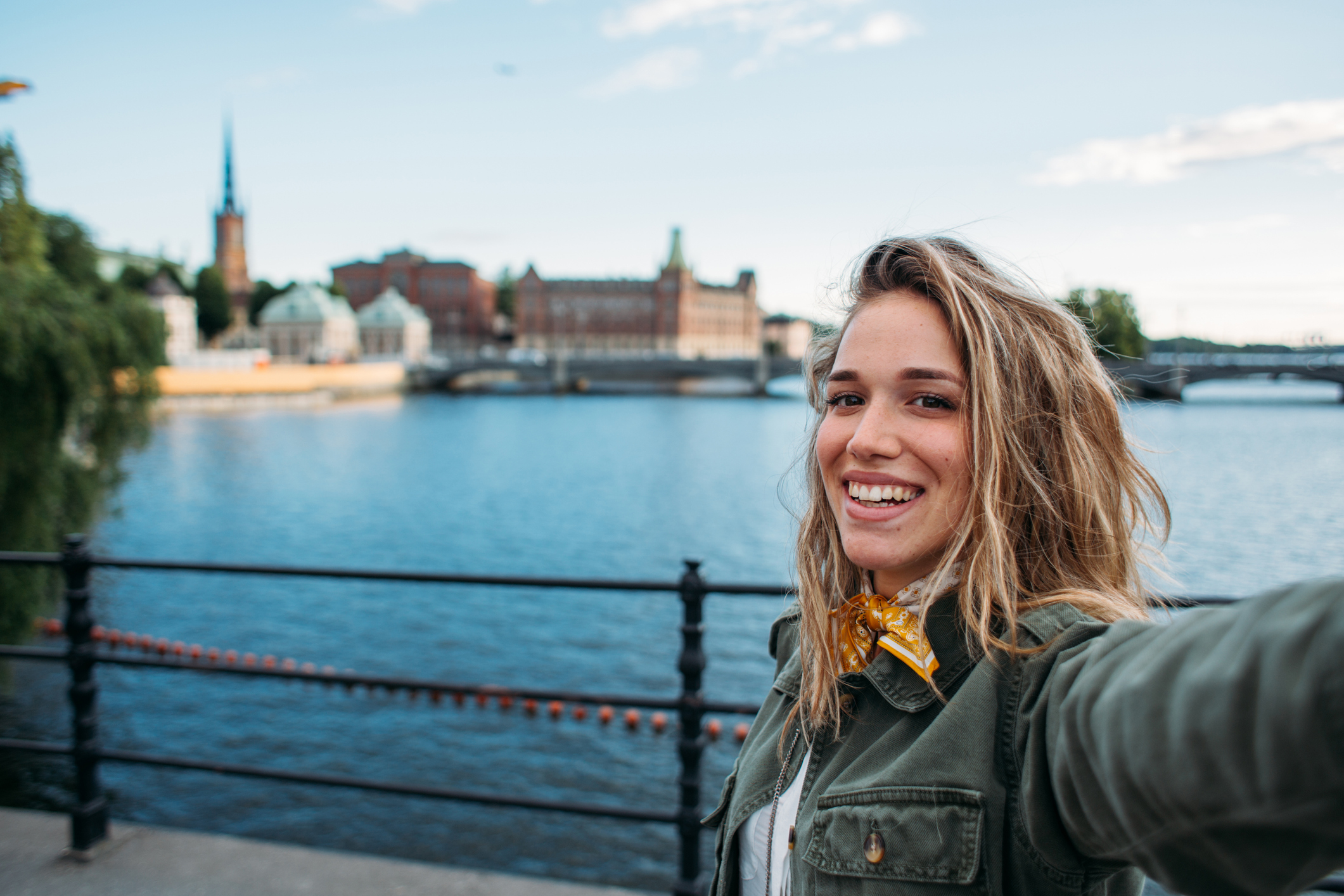 A smiling person takes a selfie by a waterfront with buildings and a spire in the background under a clear blue sky.