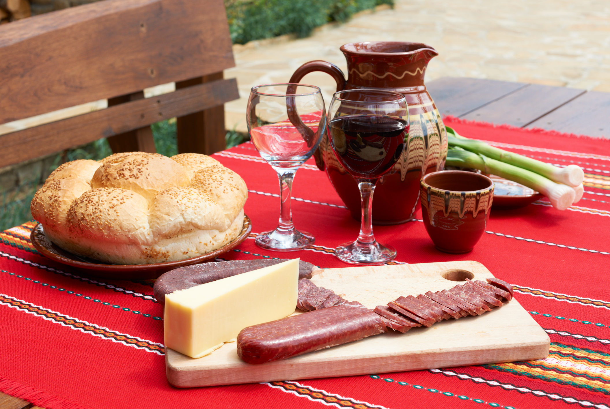 A wooden table set with bread, cheese, cured meats, wine glasses, and a decorated pitcher on a colorful tablecloth.