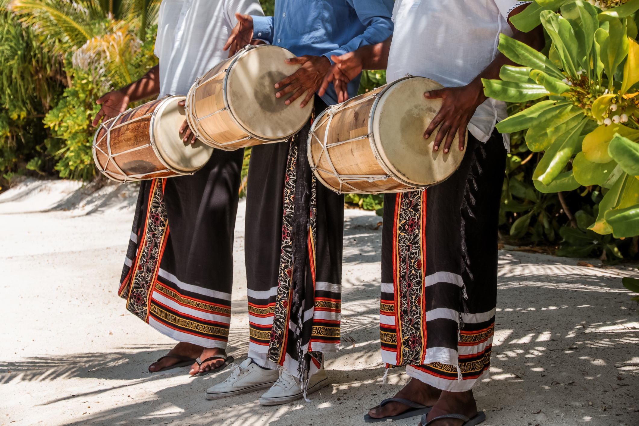Three musicians in traditional attire playing drums outdoors, surrounded by lush greenery.