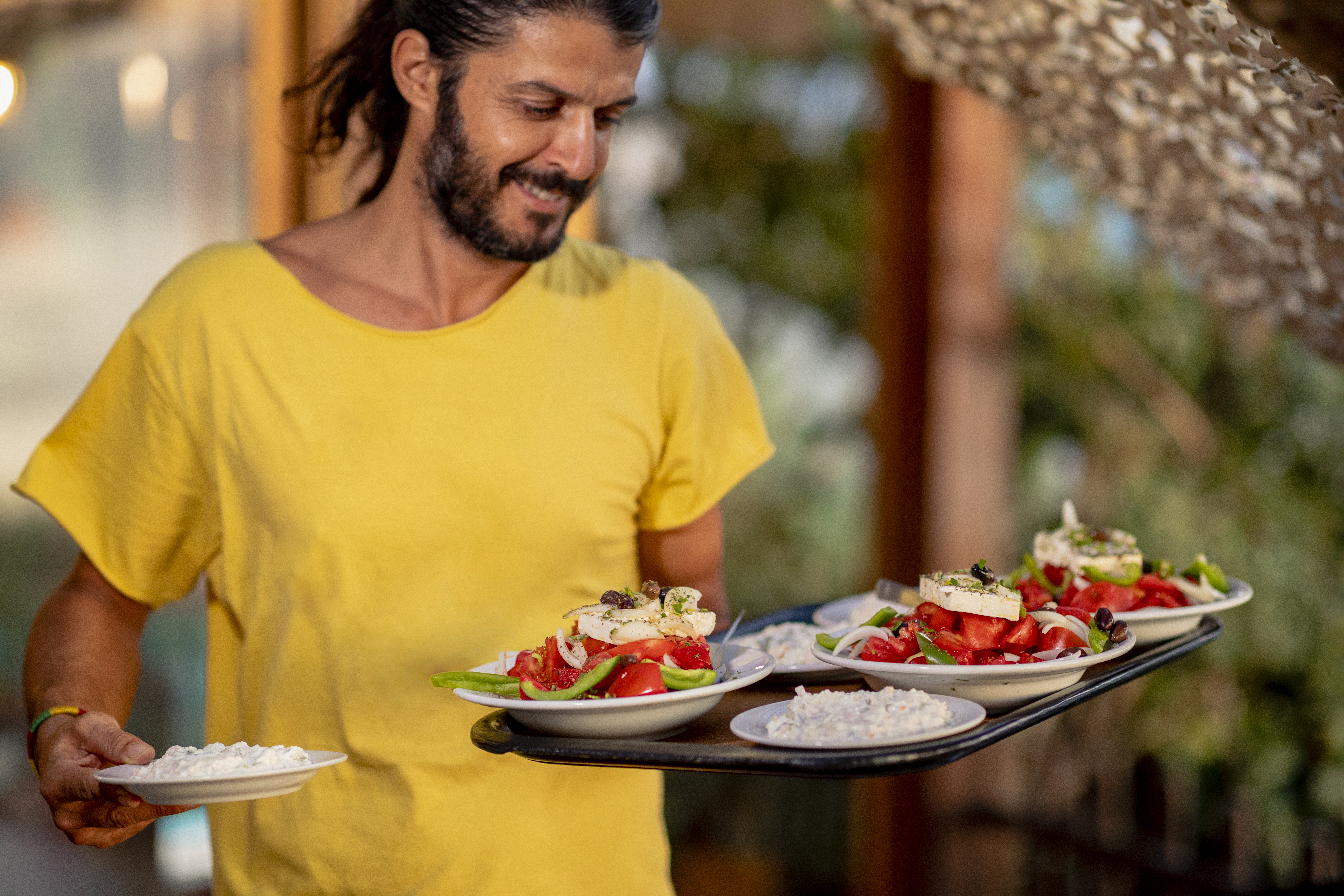 A man in a yellow shirt holds a tray with salads and a small bowl of white food, smiling in a bright setting.
