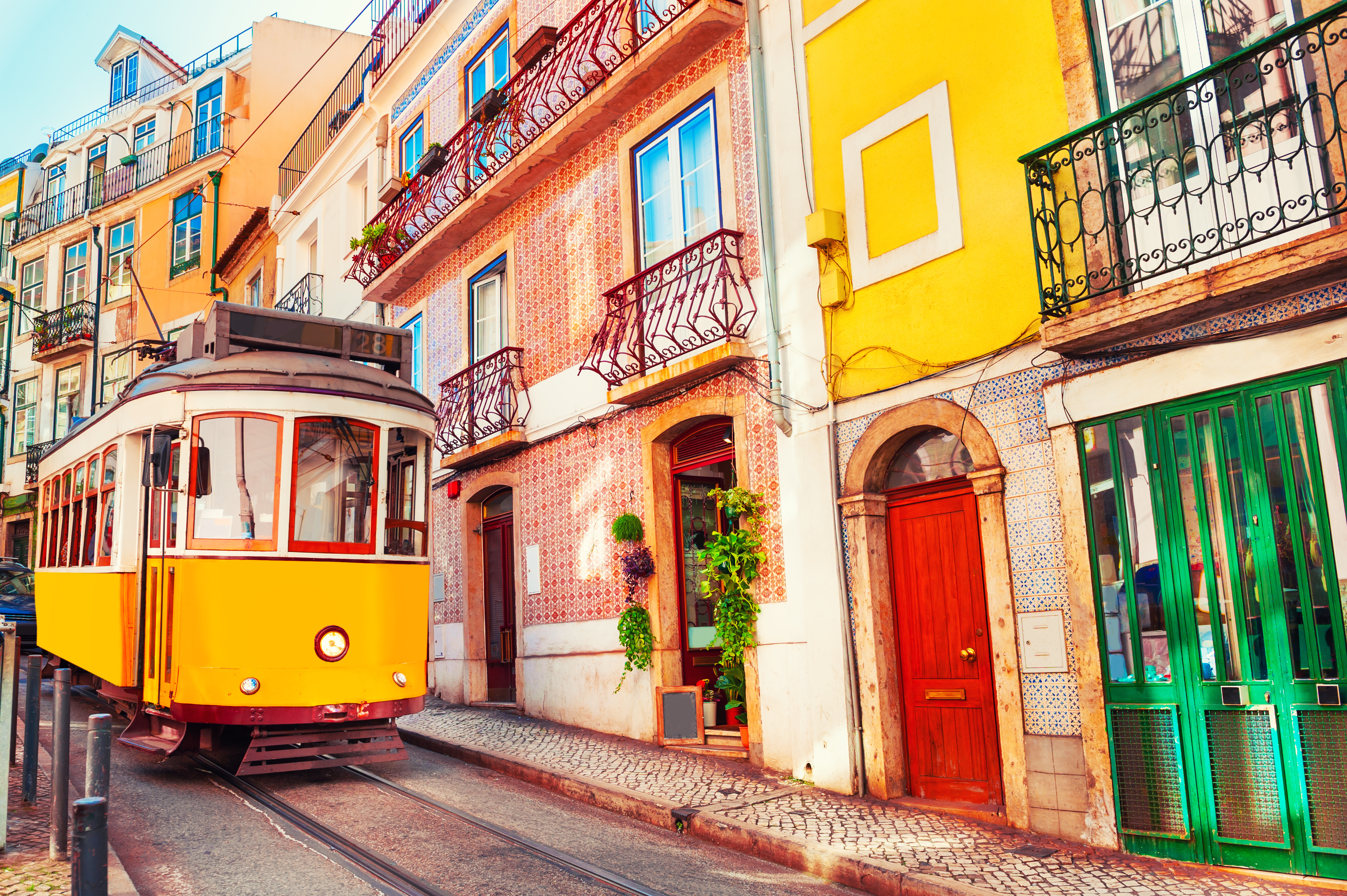 A vintage yellow tram travels along a cobblestone street lined with colorful buildings and balconies in a scenic neighborhood.