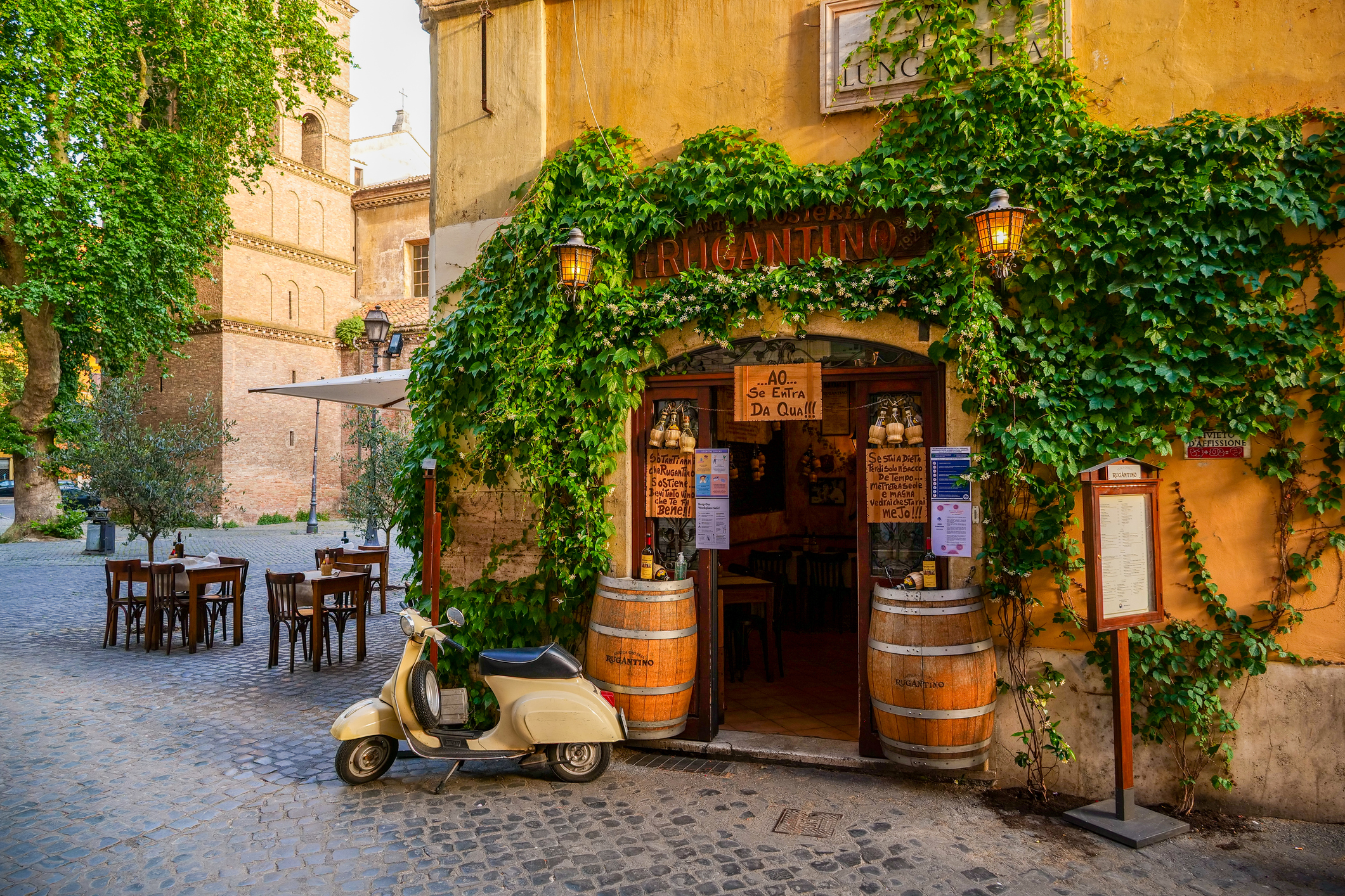 Charming outdoor café entrance with greenery, a vintage scooter, and rustic wooden tables in a cobblestone square.