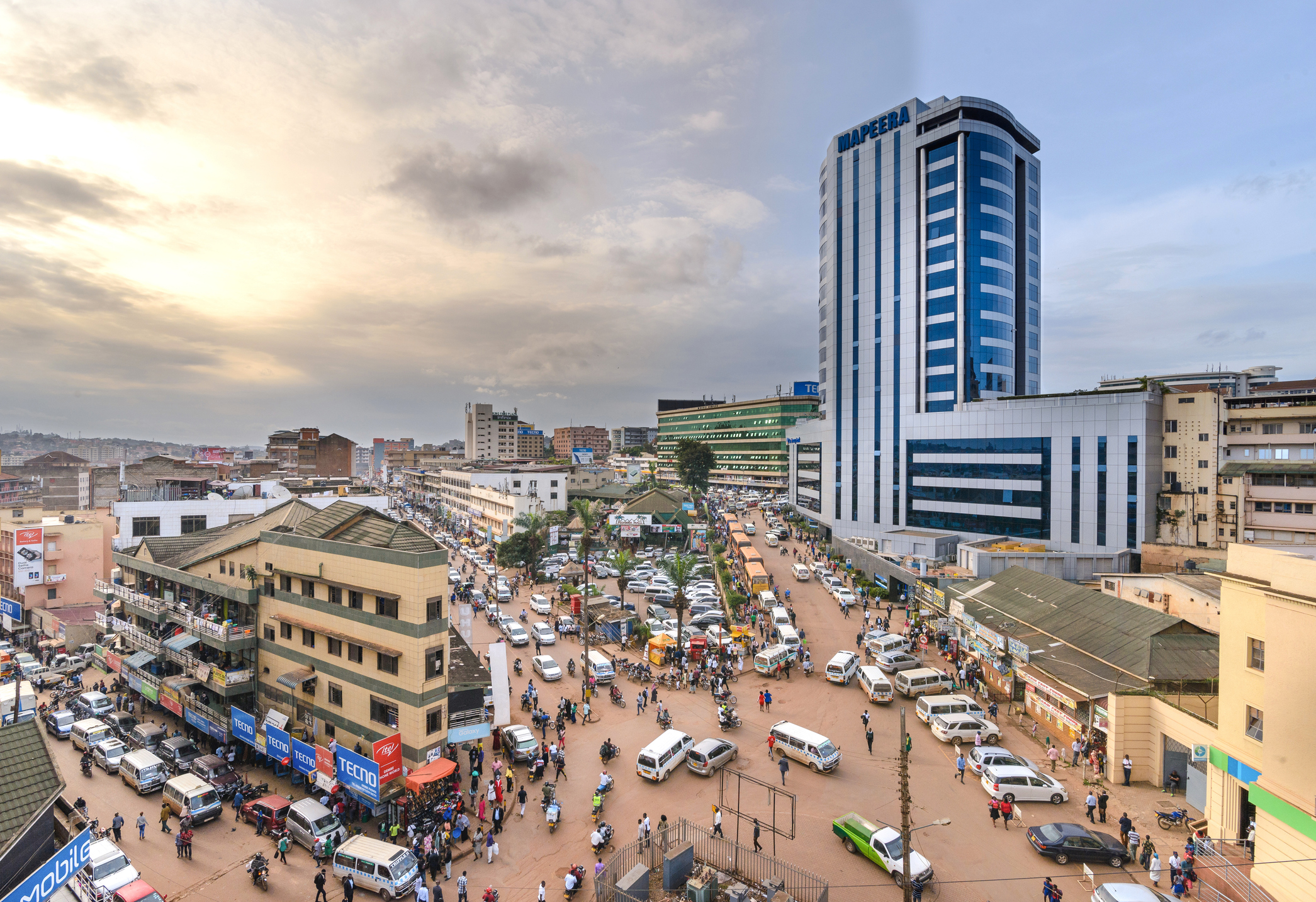 A bustling urban market scene with cars, people, and tall buildings under a cloudy sky during sunset.