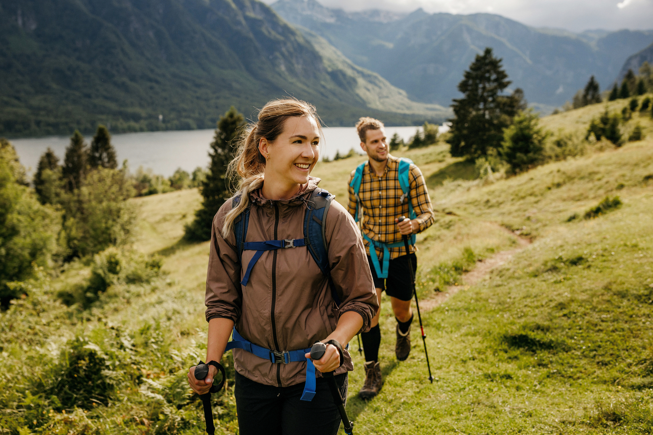 A woman and man hike on a grassy trail with mountains and a lake in the background, enjoying nature.