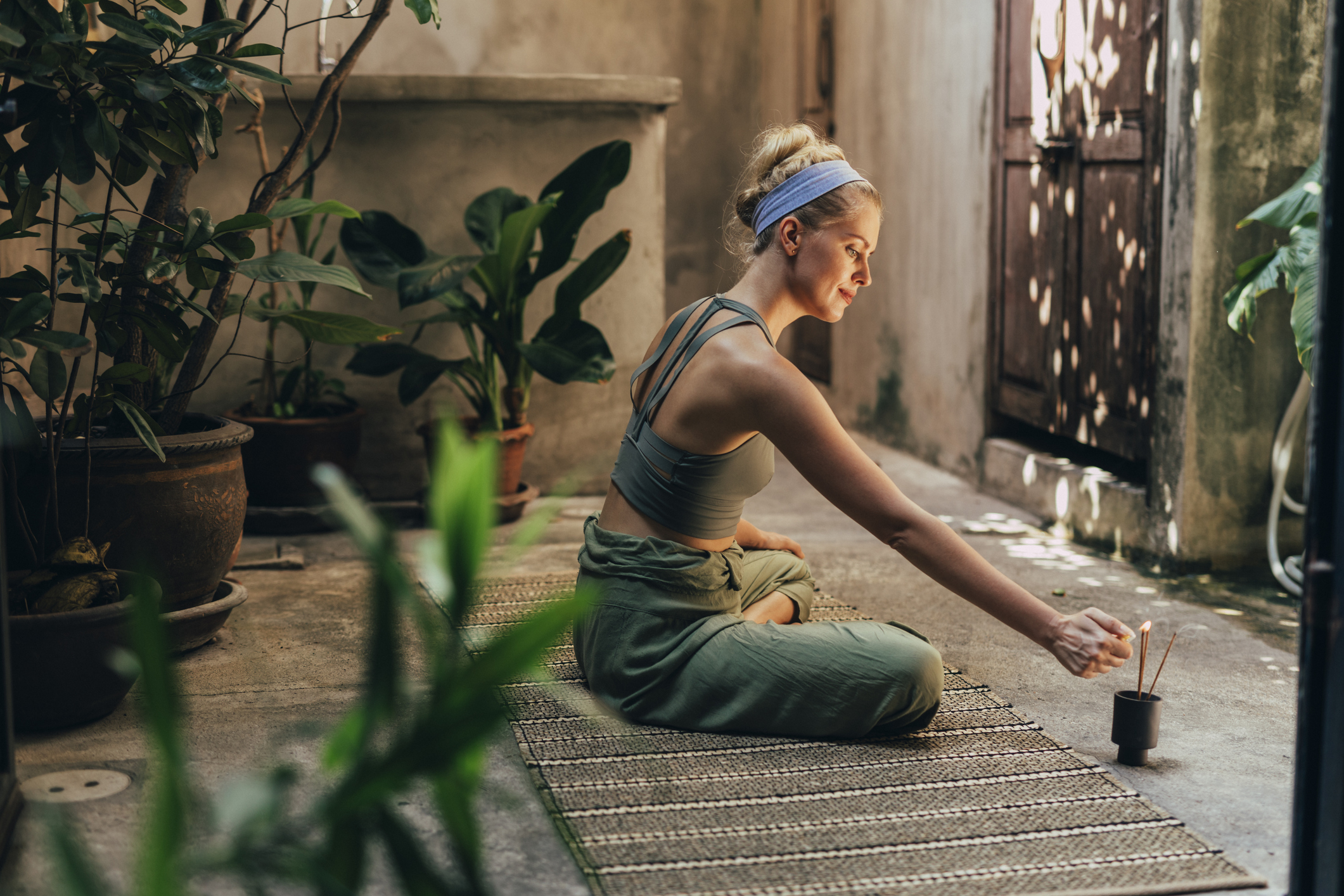 A woman sits cross-legged on the floor in a serene outdoor space, lighting an incense stick surrounded by plants.