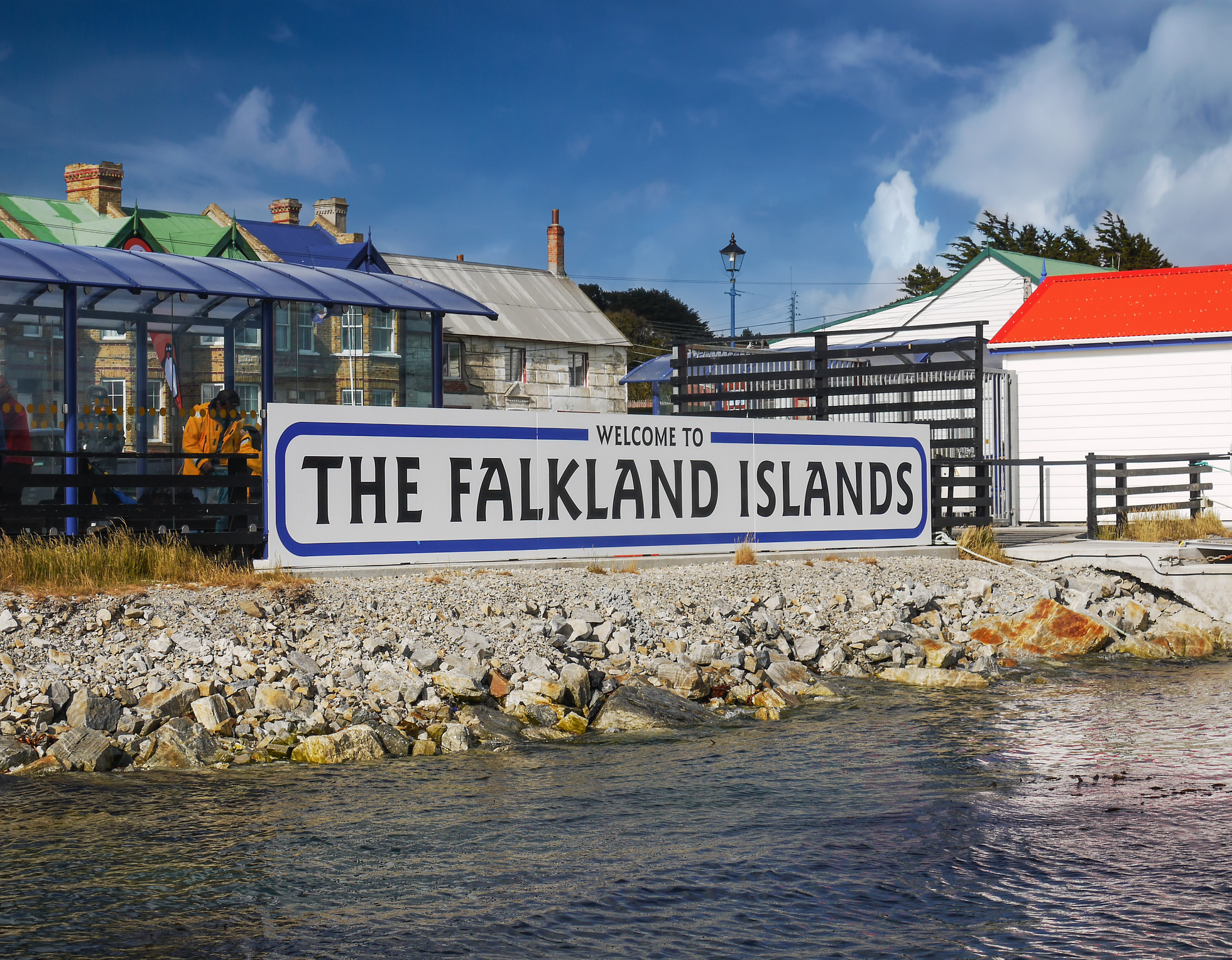 Welcome sign for the Falkland Islands near a waterfront, with buildings in the background and a cloudy sky.