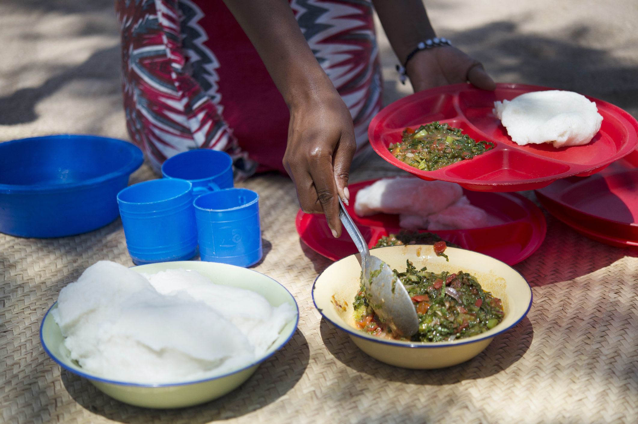 A person serves food from a bowl onto colorful plates, with cups nearby, on a textured surface outdoors.