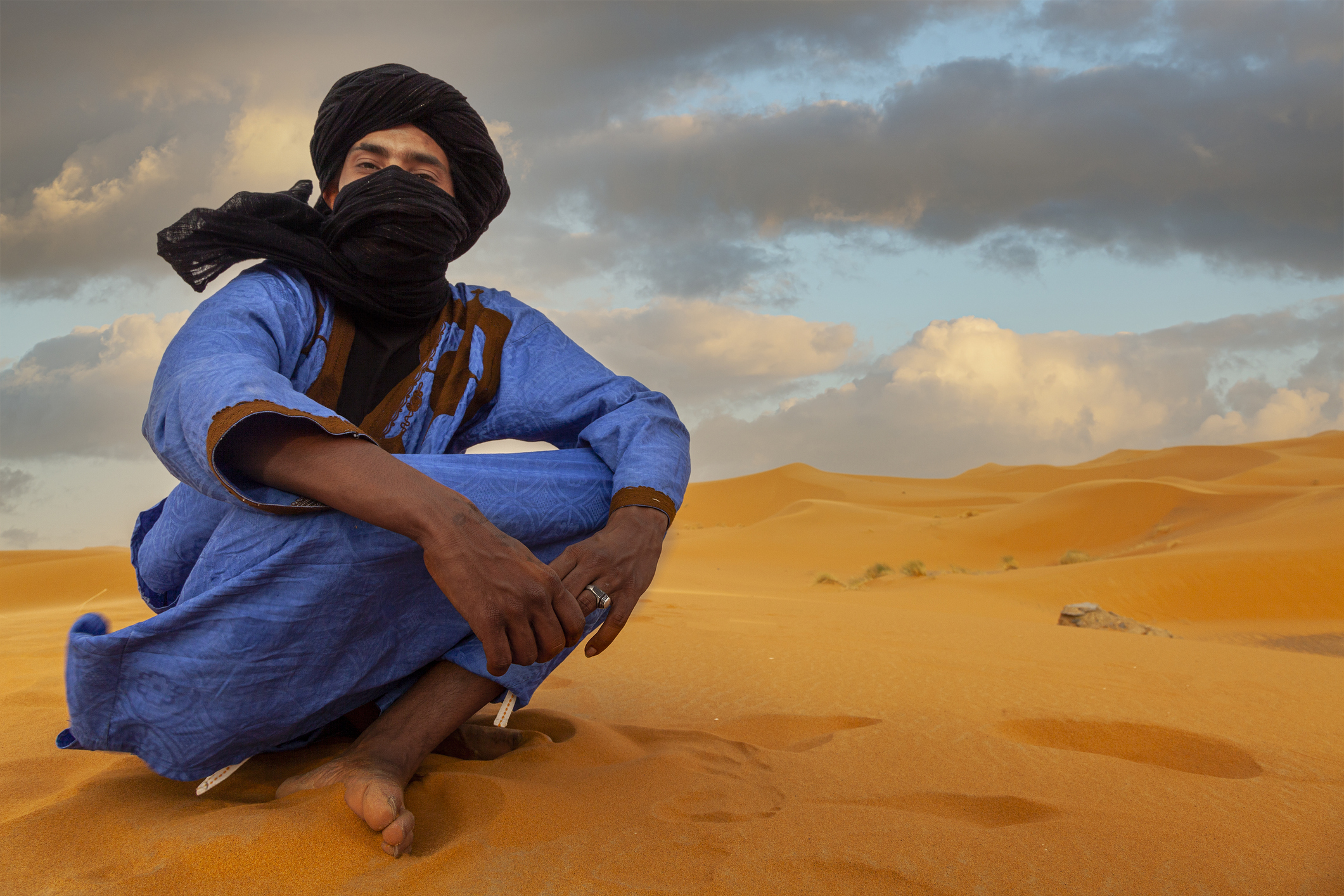 A person in a blue robe and black scarf sits on golden sand dunes under a cloudy sky.