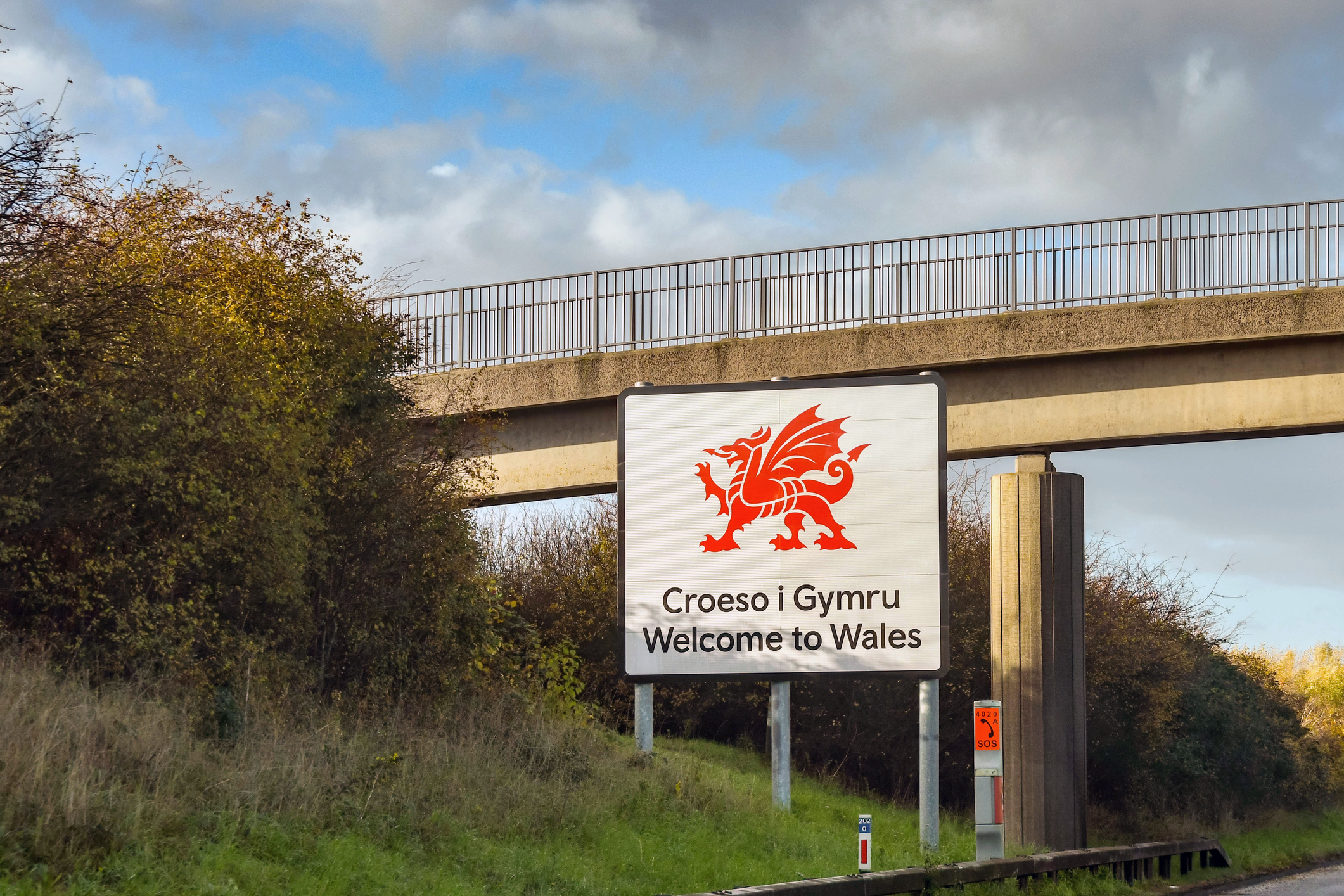 Sign welcoming visitors to Wales with a red dragon emblem, bordered by greenery and a bridge overhead.