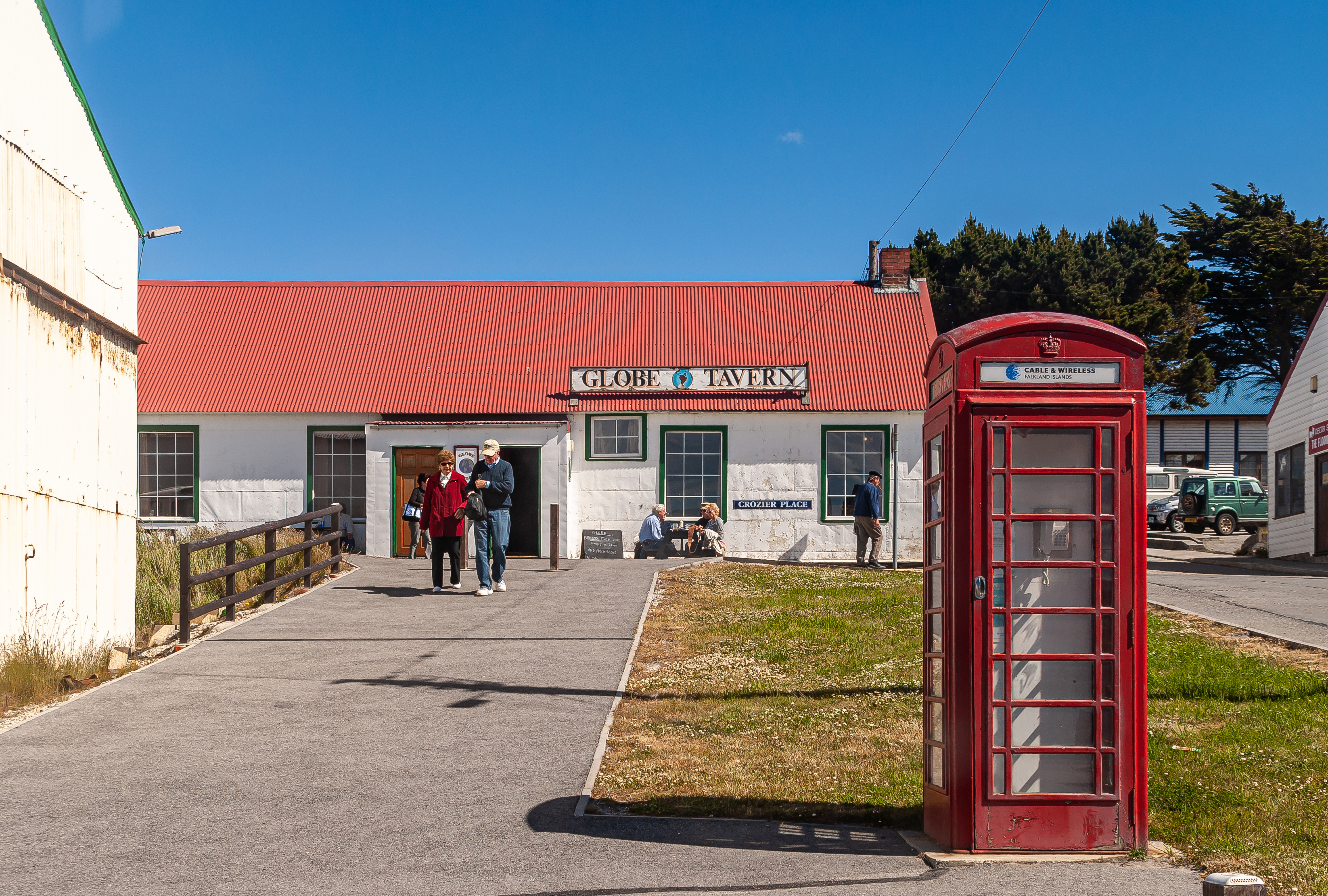 A red telephone box stands in front of the Globe Tavern, with people walking nearby and a clear blue sky above.