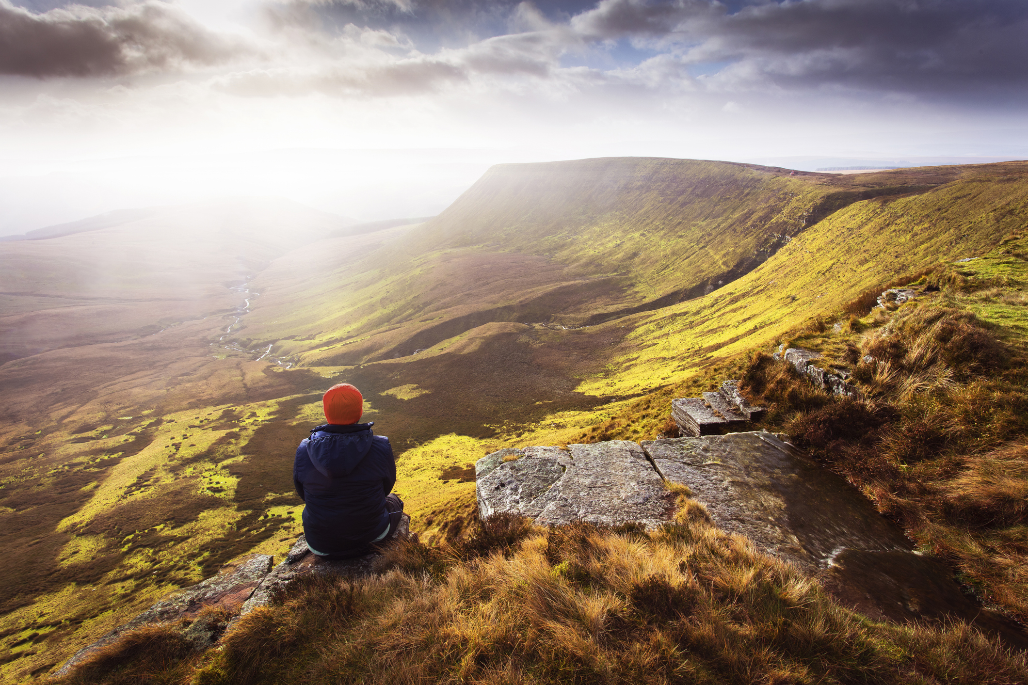 Person sitting on a rocky ledge, gazing at a vast green valley under a cloudy sky.