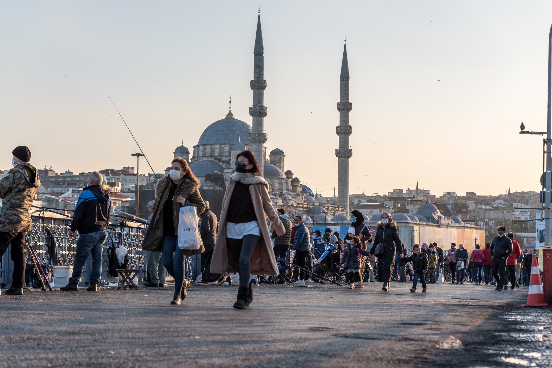 Crowds of people walk along a busy bridge with the skyline and mosque minarets in the background at sunset.