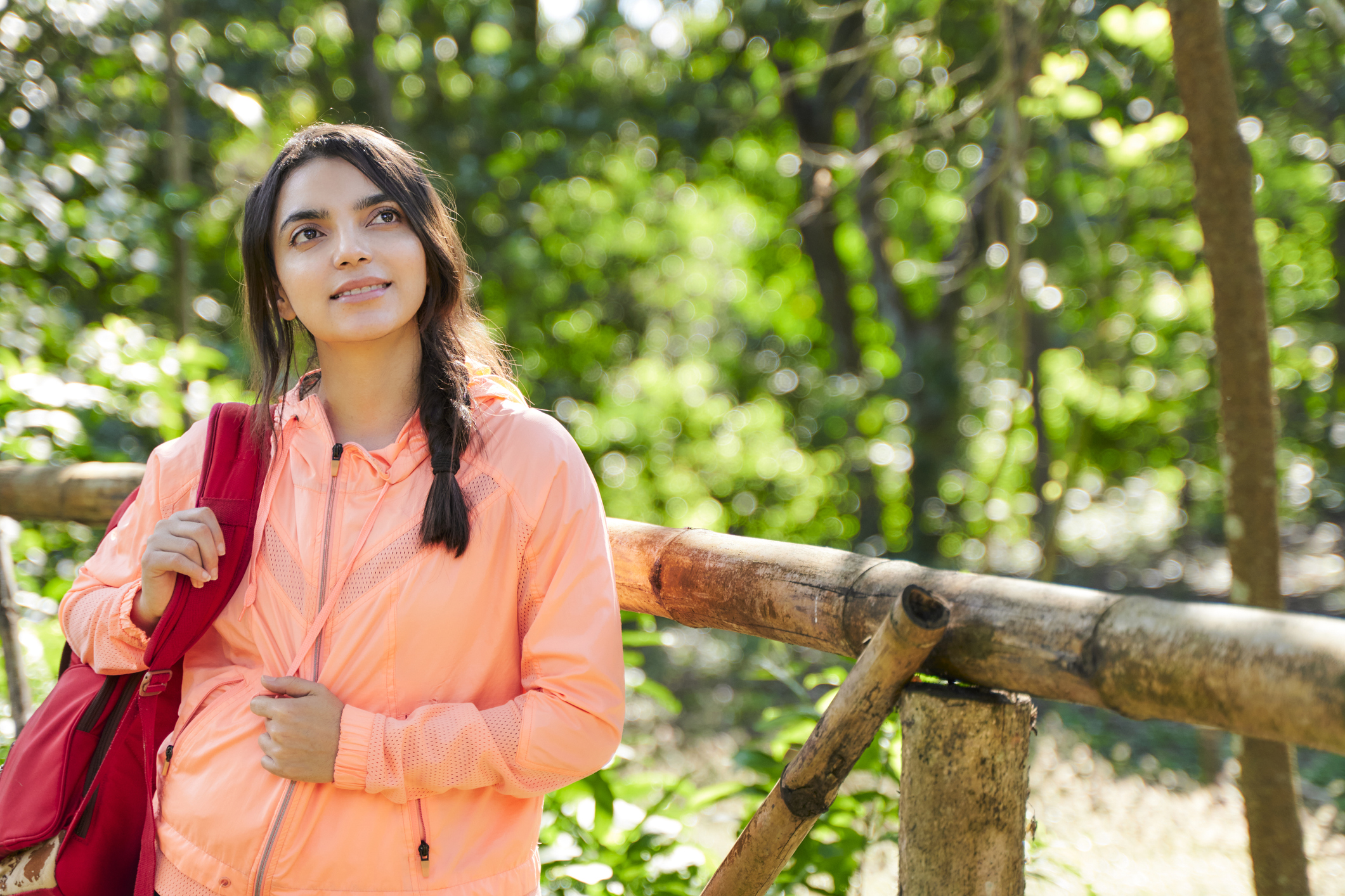 A young woman in an orange jacket stands by a wooden fence, looking thoughtfully into the distance in a lush forest.