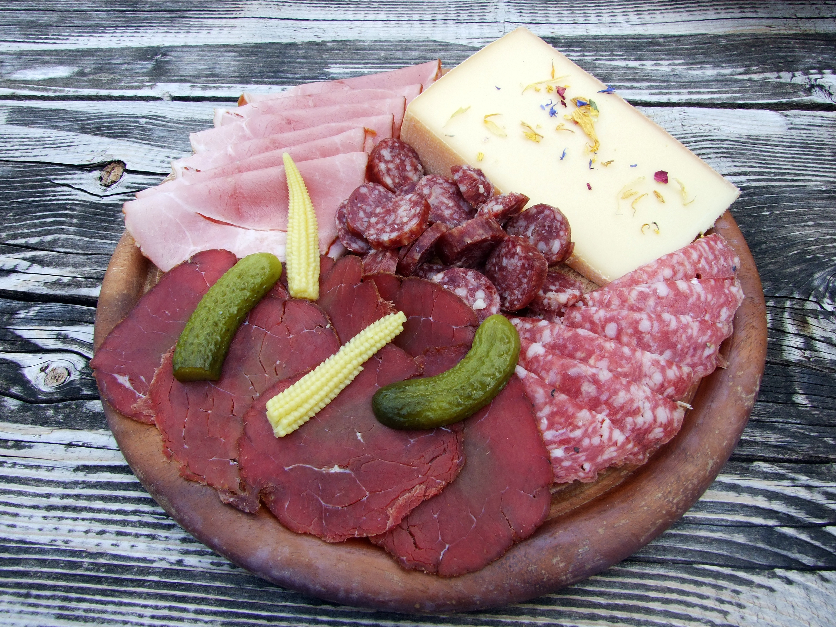 A wooden platter with assorted meats, cheeses, and pickles, including ham, salami, and baby corn.