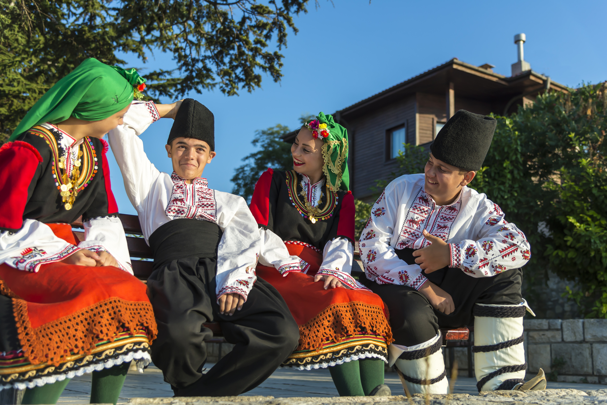 Four young people in traditional Bulgarian clothing pose together outdoors, smiling and engaging with each other.