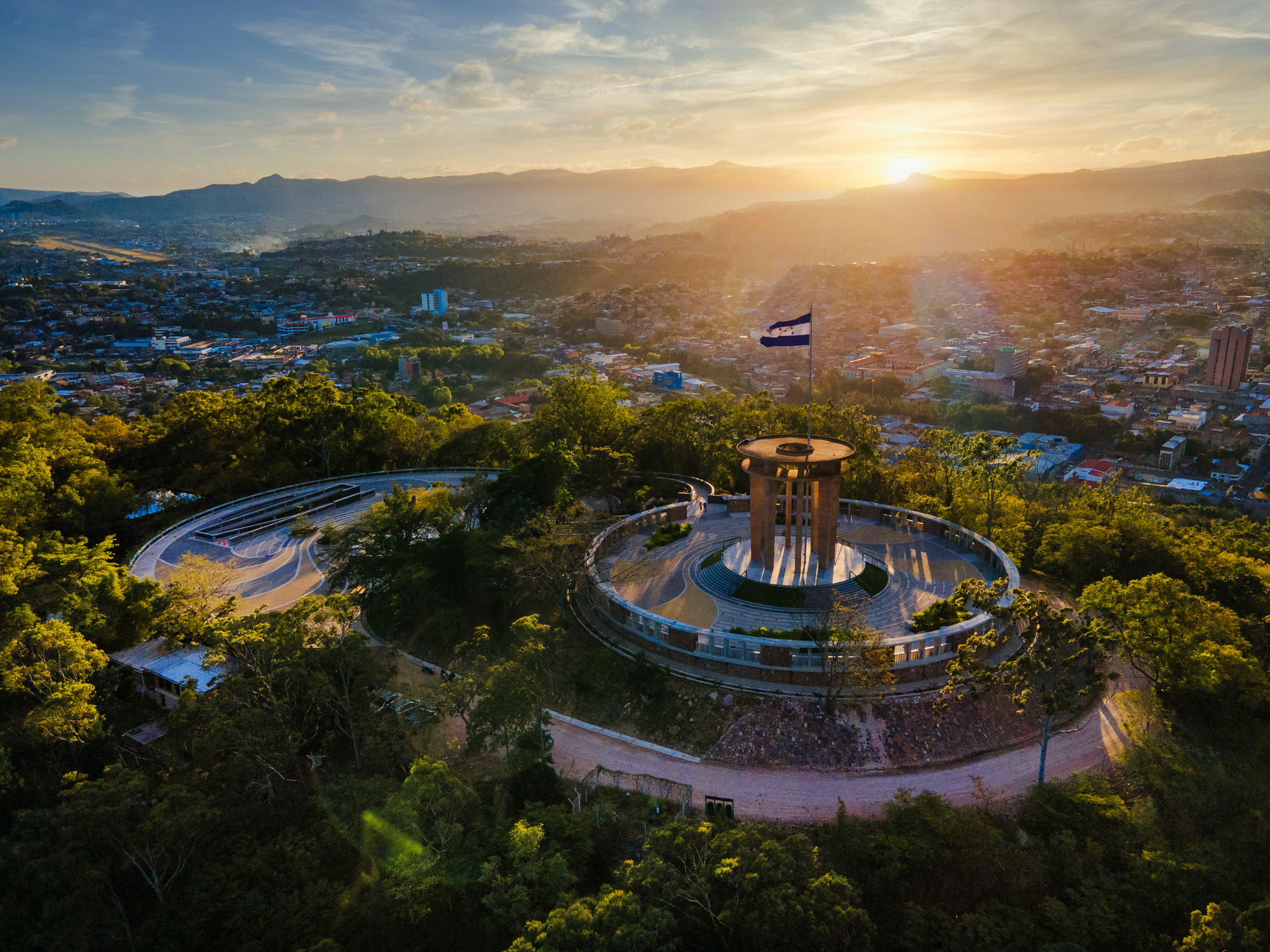 Aerial view of a scenic lookout with circular paths, a monument, and a flag, surrounded by trees and a city backdrop at sunset.