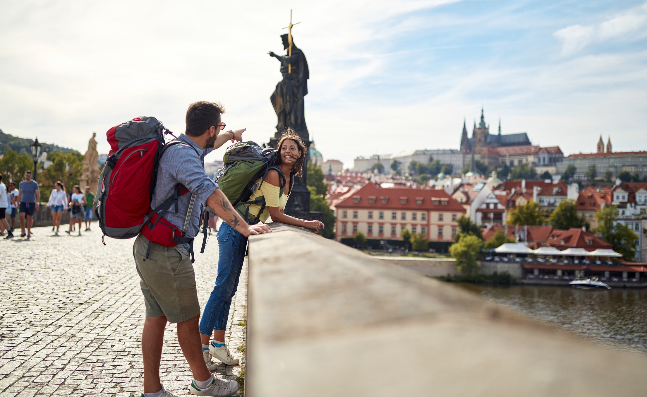 Two travelers with backpacks enjoy the view from a bridge, with historic buildings and a statue in the background.