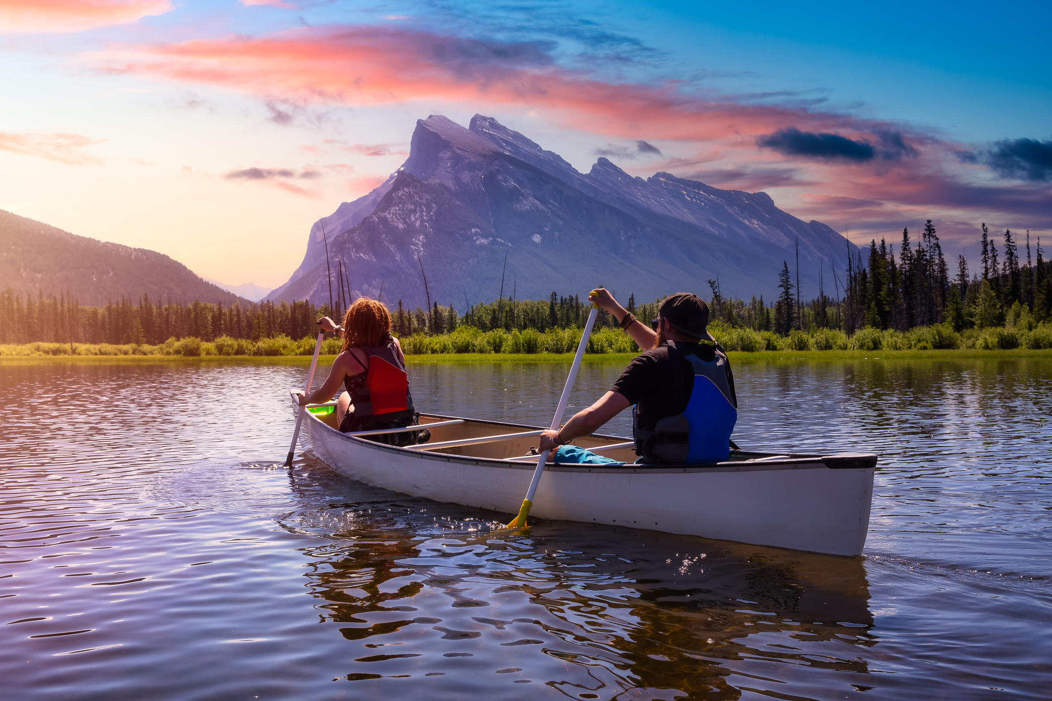 Two people paddle a canoe on a calm lake, with mountains and colorful sunset in the background.