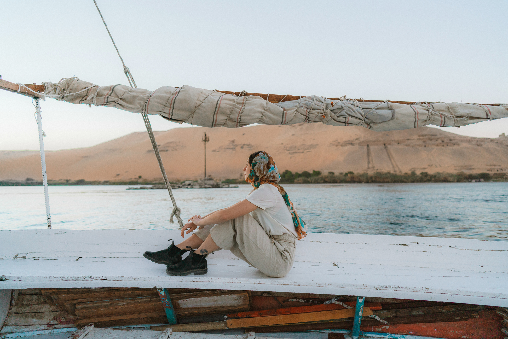 Person sitting on a boat, wearing a scarf, with desert hills and water in the background during sunset.