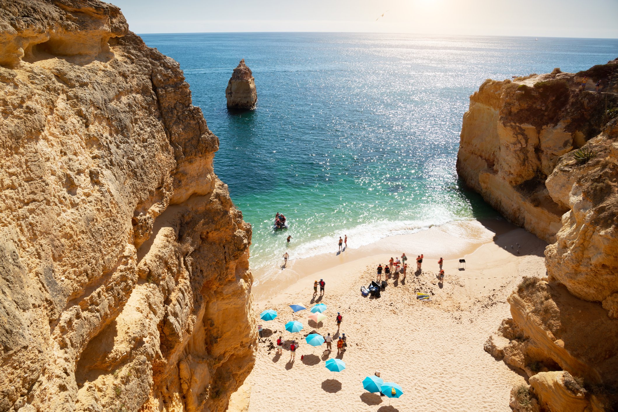 A sunlit beach with people near turquoise water, surrounded by rocky cliffs and colorful umbrellas.
