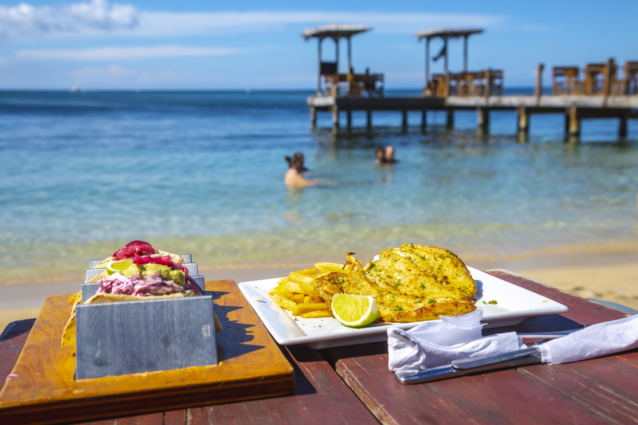 A beachside table displays two dishes: a colorful platter and a grilled fish dish, with people swimming in the background.