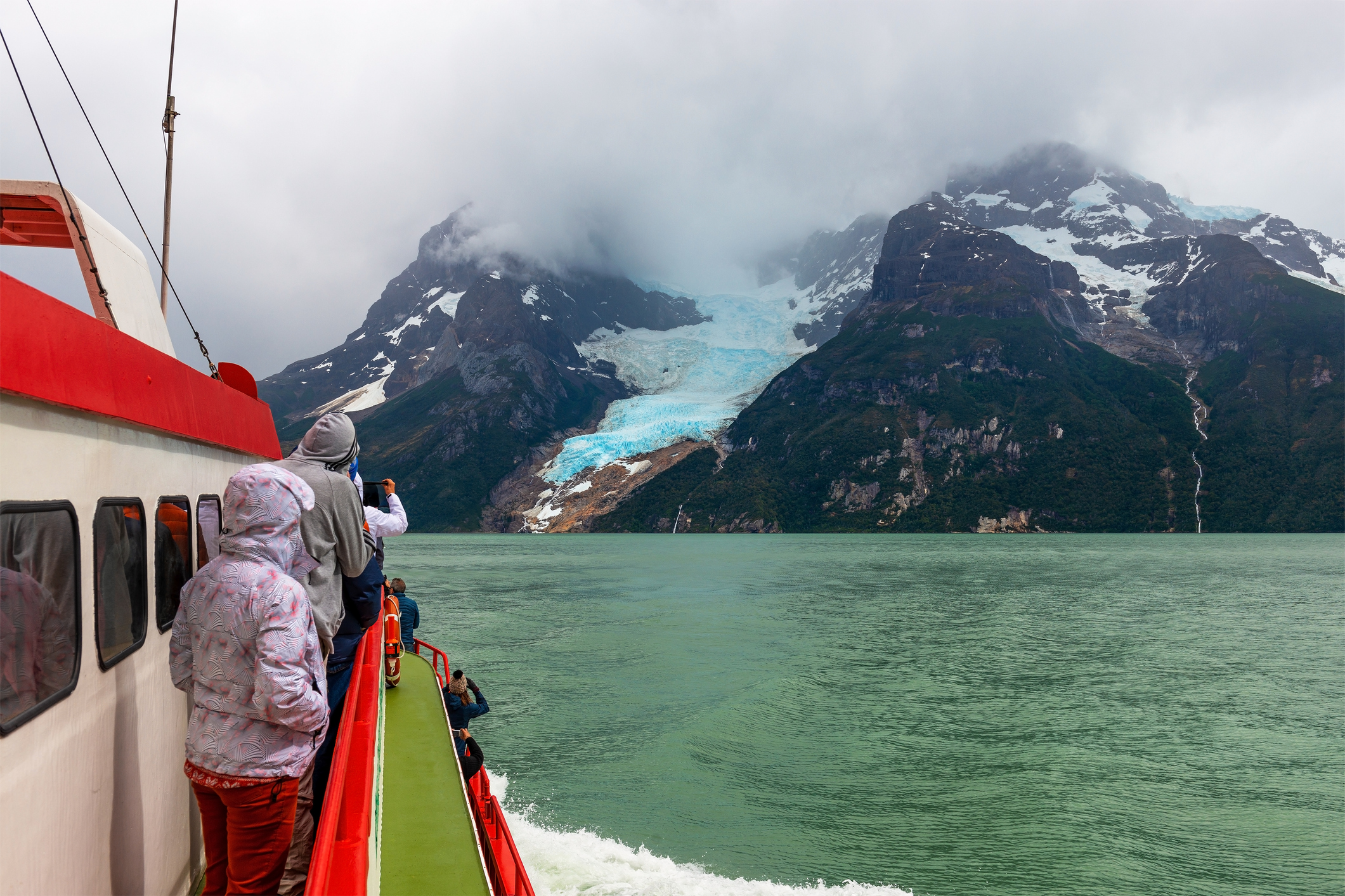 Passengers on a boat view a glacier and mountains under a cloudy sky, with water visible in the foreground.