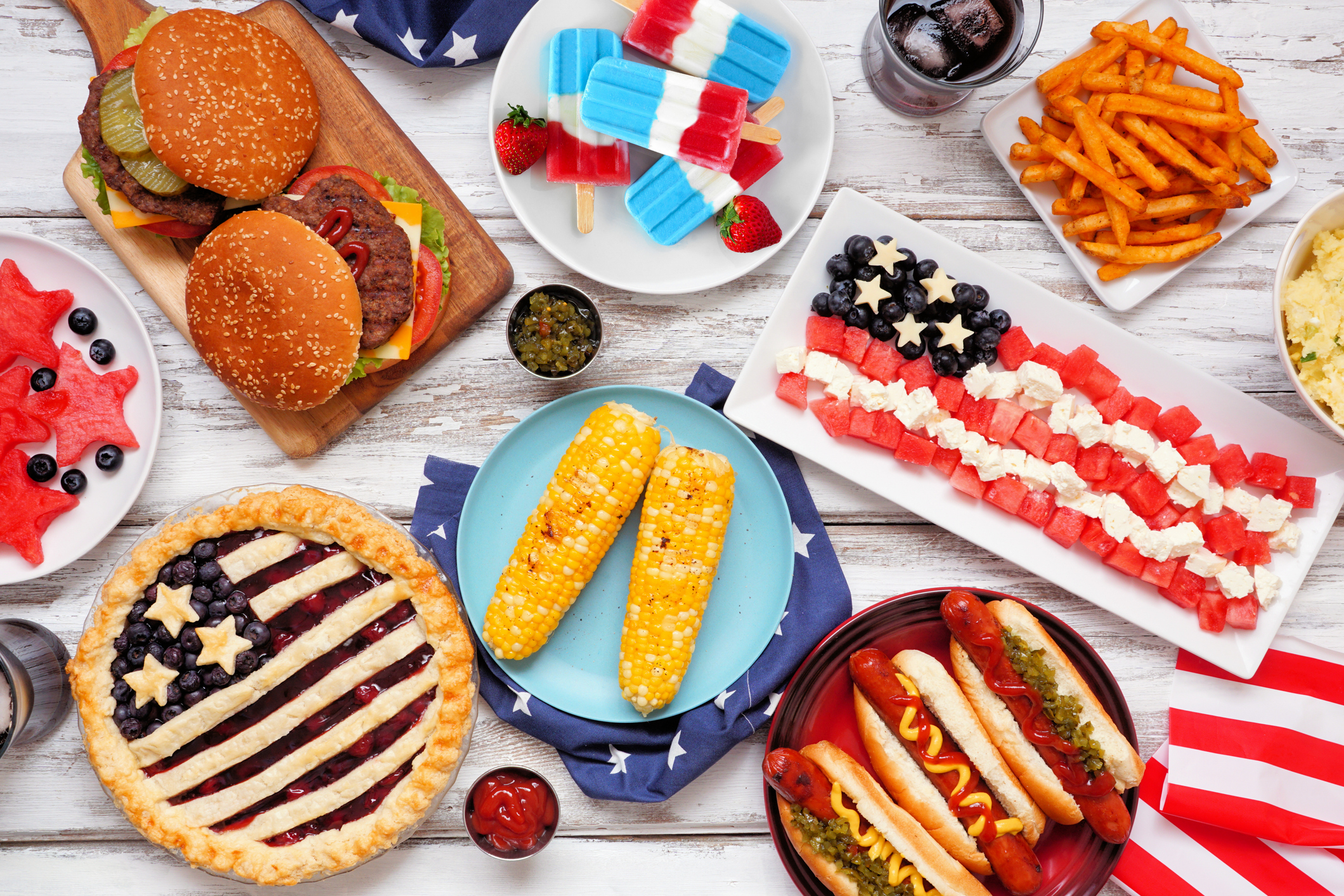 A variety of festive foods including burgers, hot dogs, watermelon, corn, and desserts arranged in a patriotic theme.