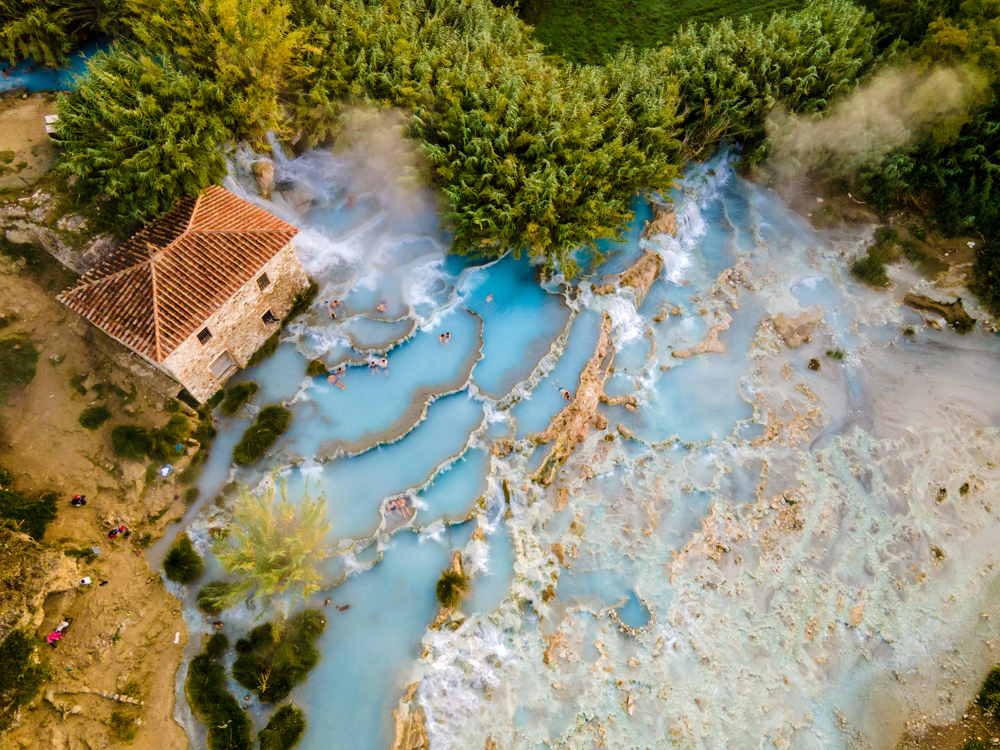 Aerial view of a rustic house near natural hot springs with blue water and lush greenery surrounding the area.