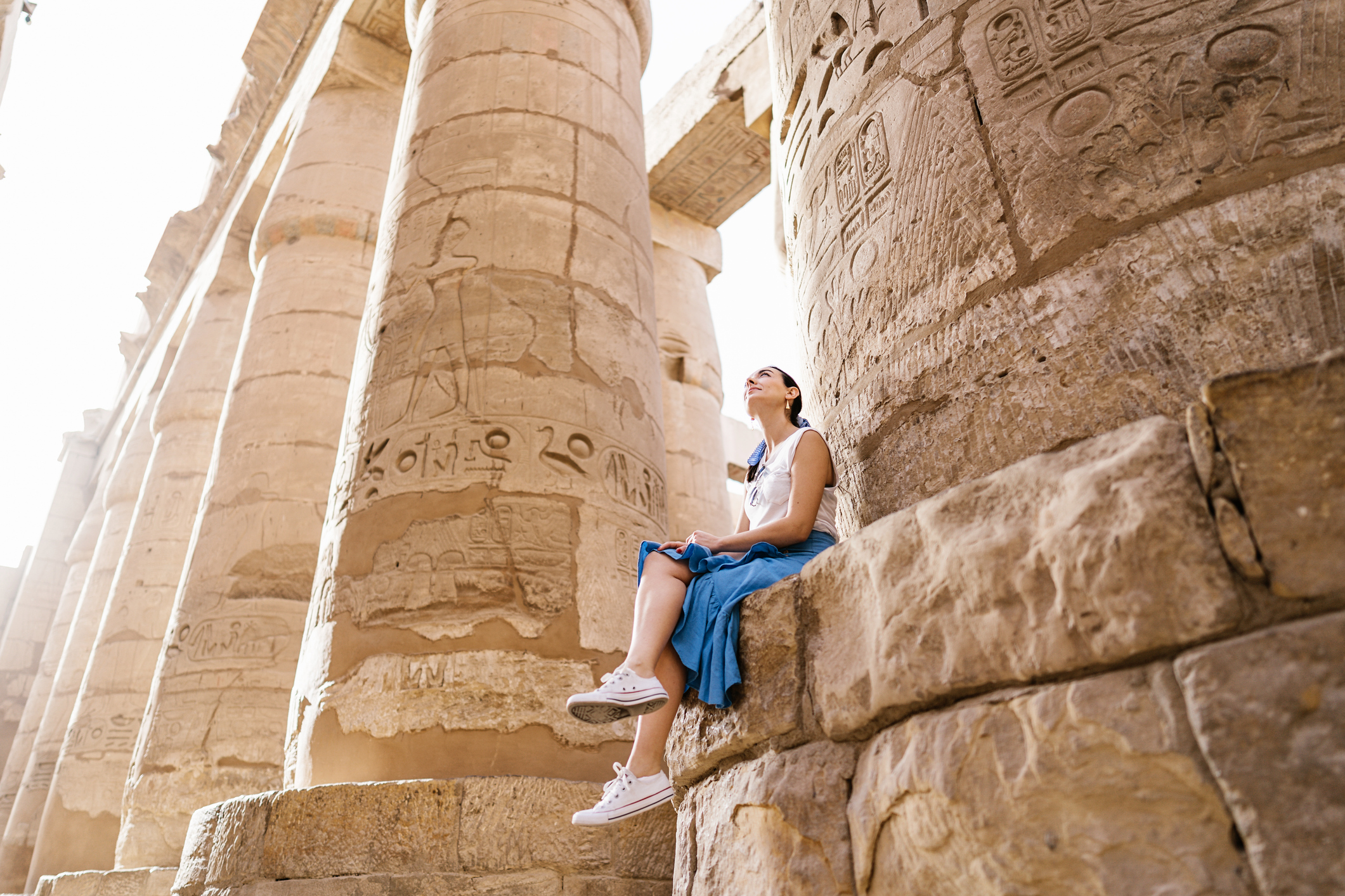 Person sitting on a stone block beside tall, ancient columns with hieroglyphics in a sunlit temple.