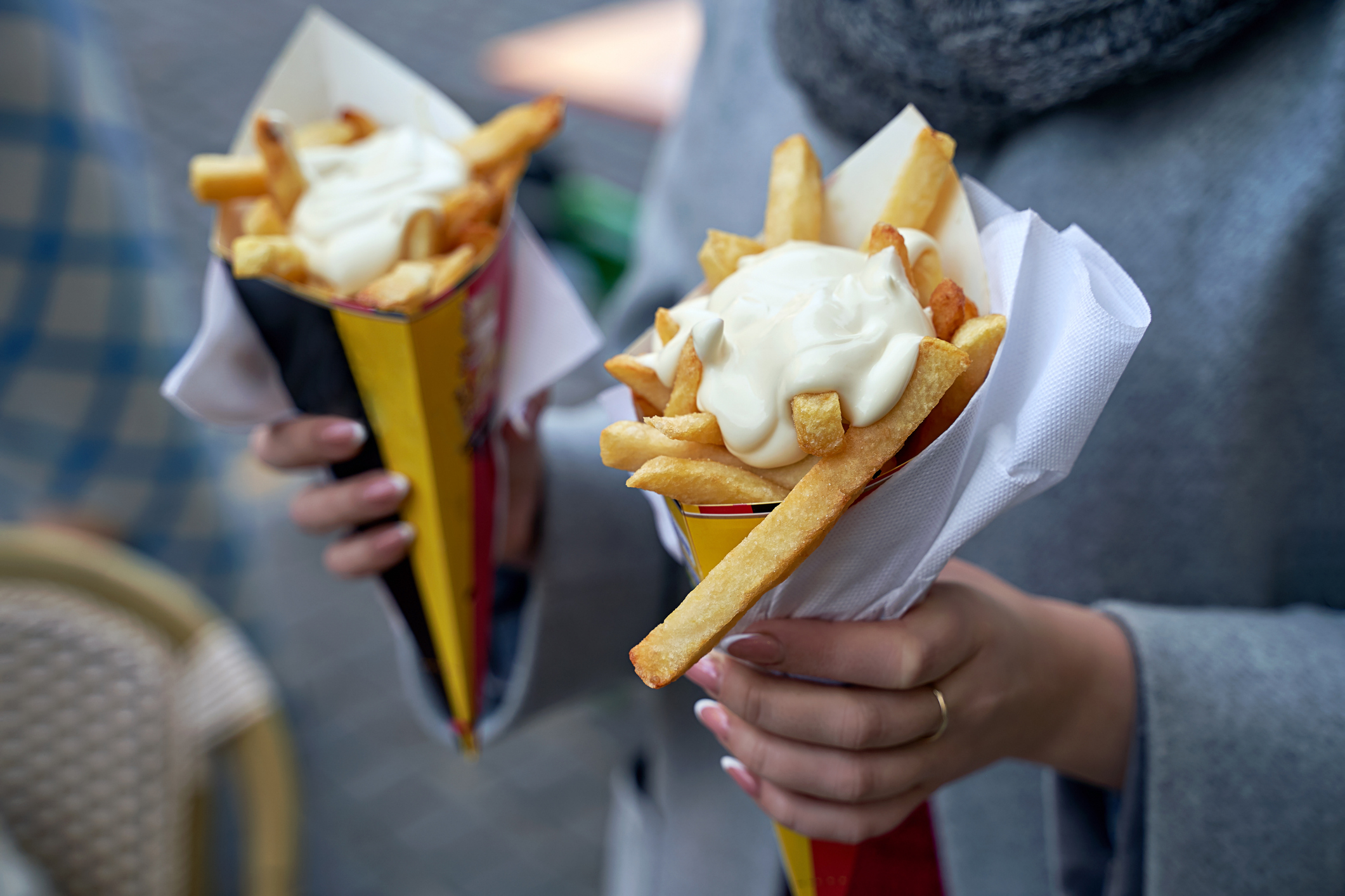 Two hands holding cones of fries topped with sauce, outdoors in a casual setting.