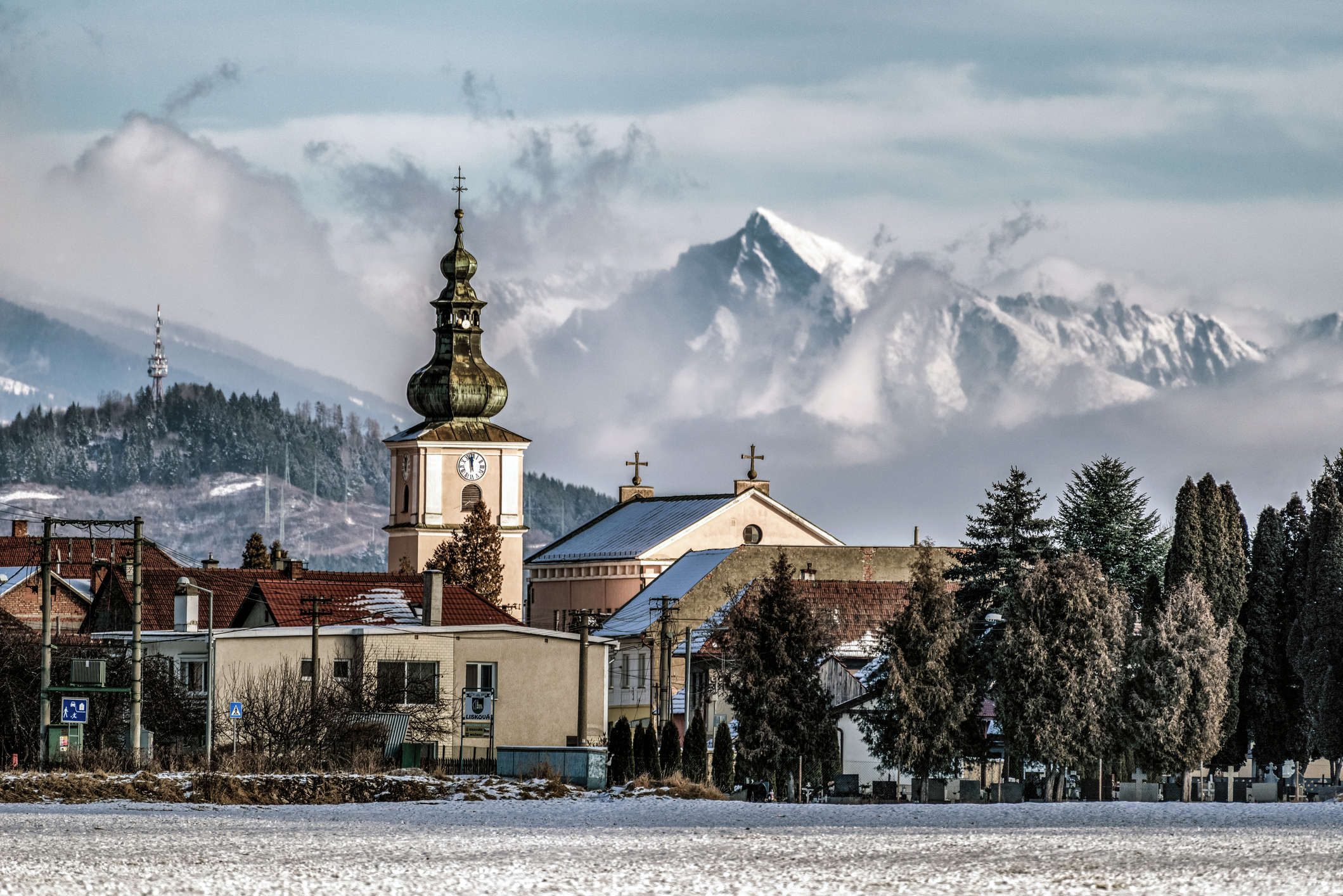 A snowy landscape with a church tower and mountains in the background, under a cloudy sky.