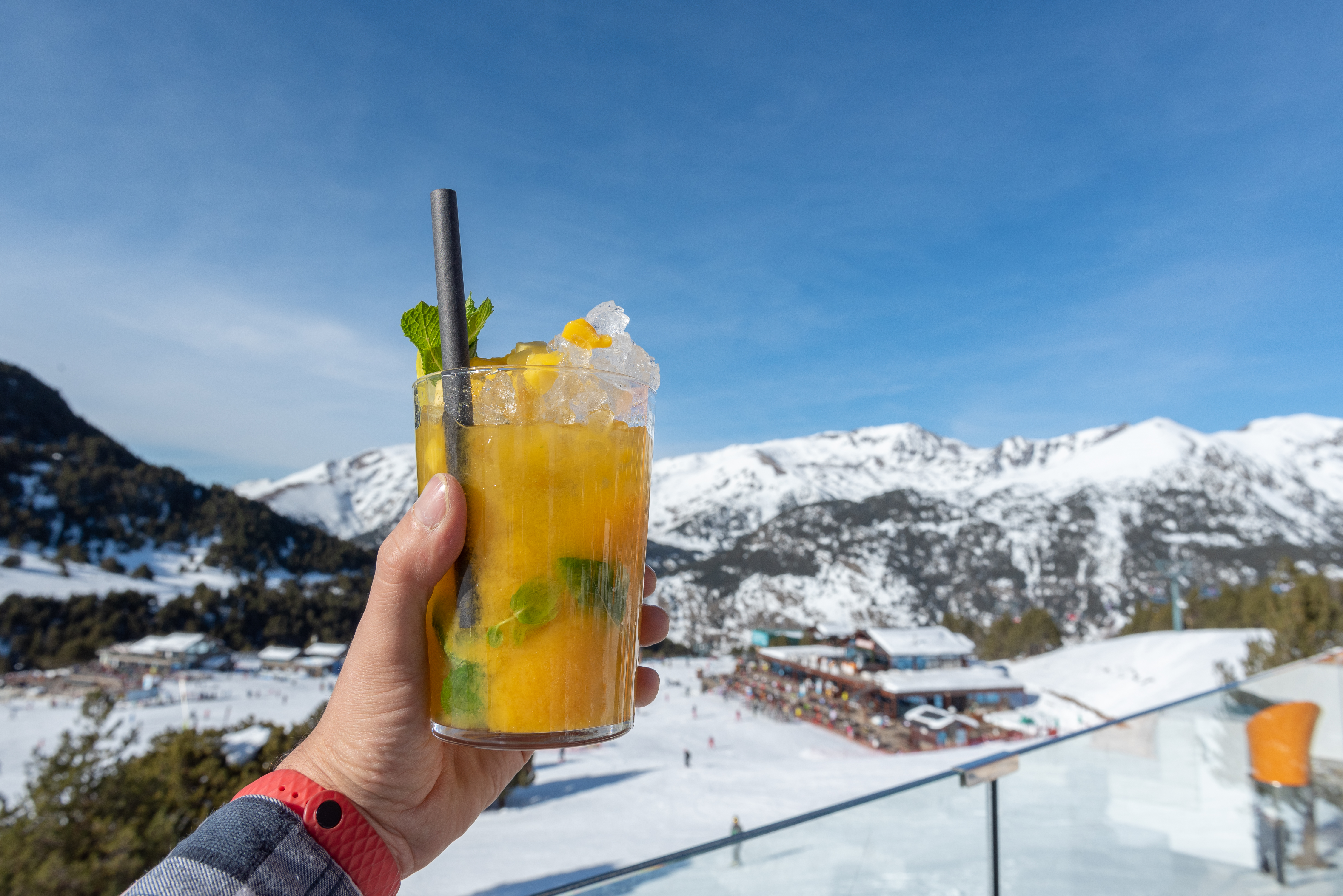 A hand holds a glass of orange cocktail with mint, set against a snowy mountain backdrop under a clear blue sky.