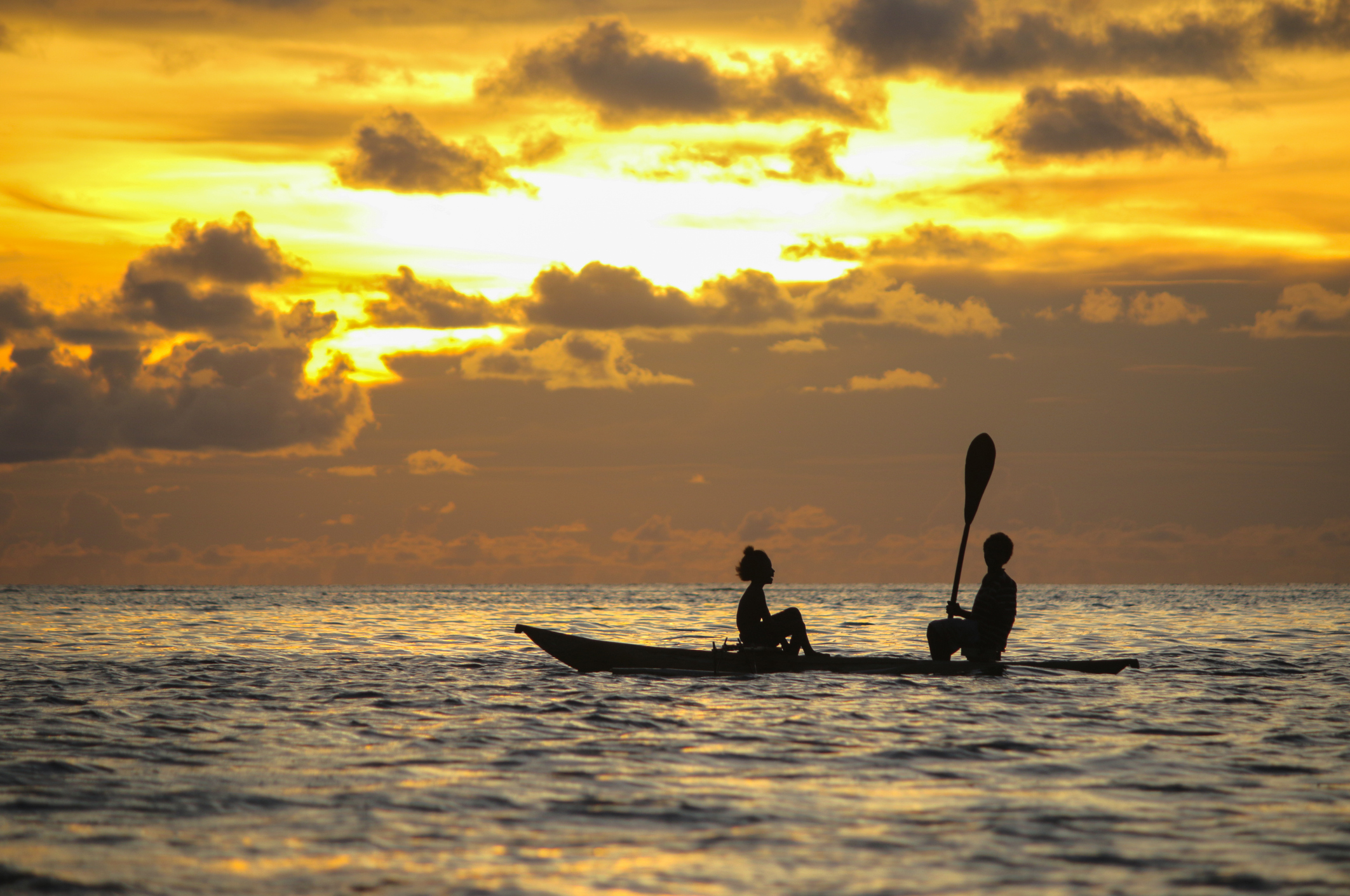Two people sit in a kayak at sunset, silhouetted against a colorful sky with clouds reflecting in the water.