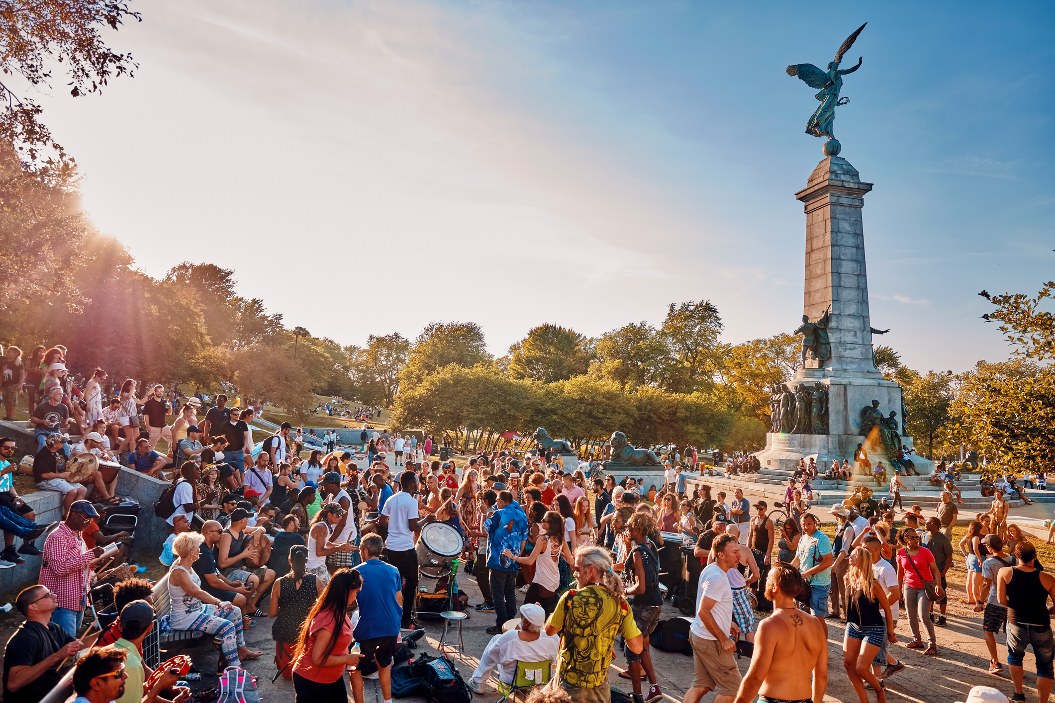 A crowded outdoor gathering near a monument with a statue, surrounded by trees and a lively atmosphere.