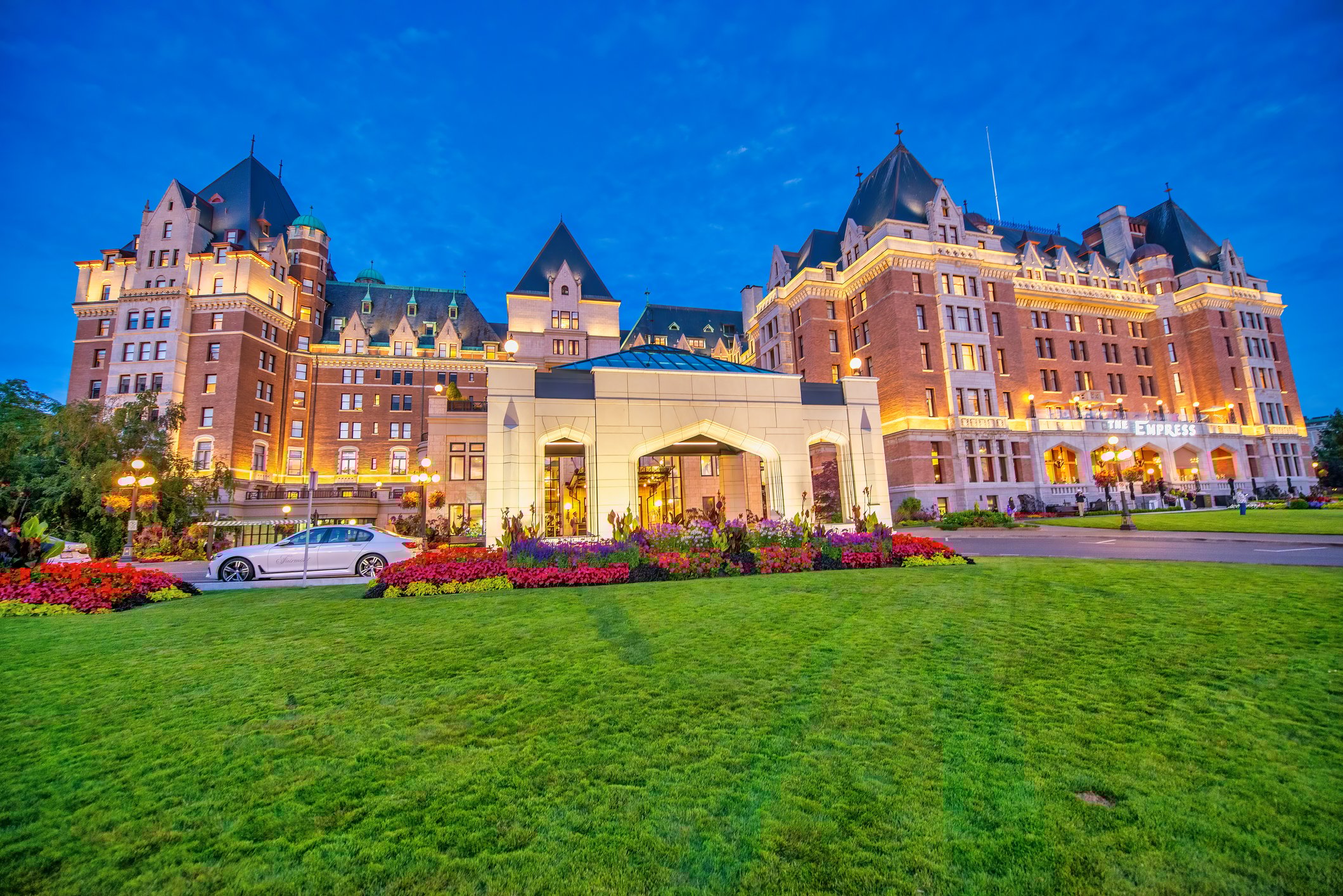 Historic hotel with a grand facade, vibrant gardens, and evening lights under a blue sky.