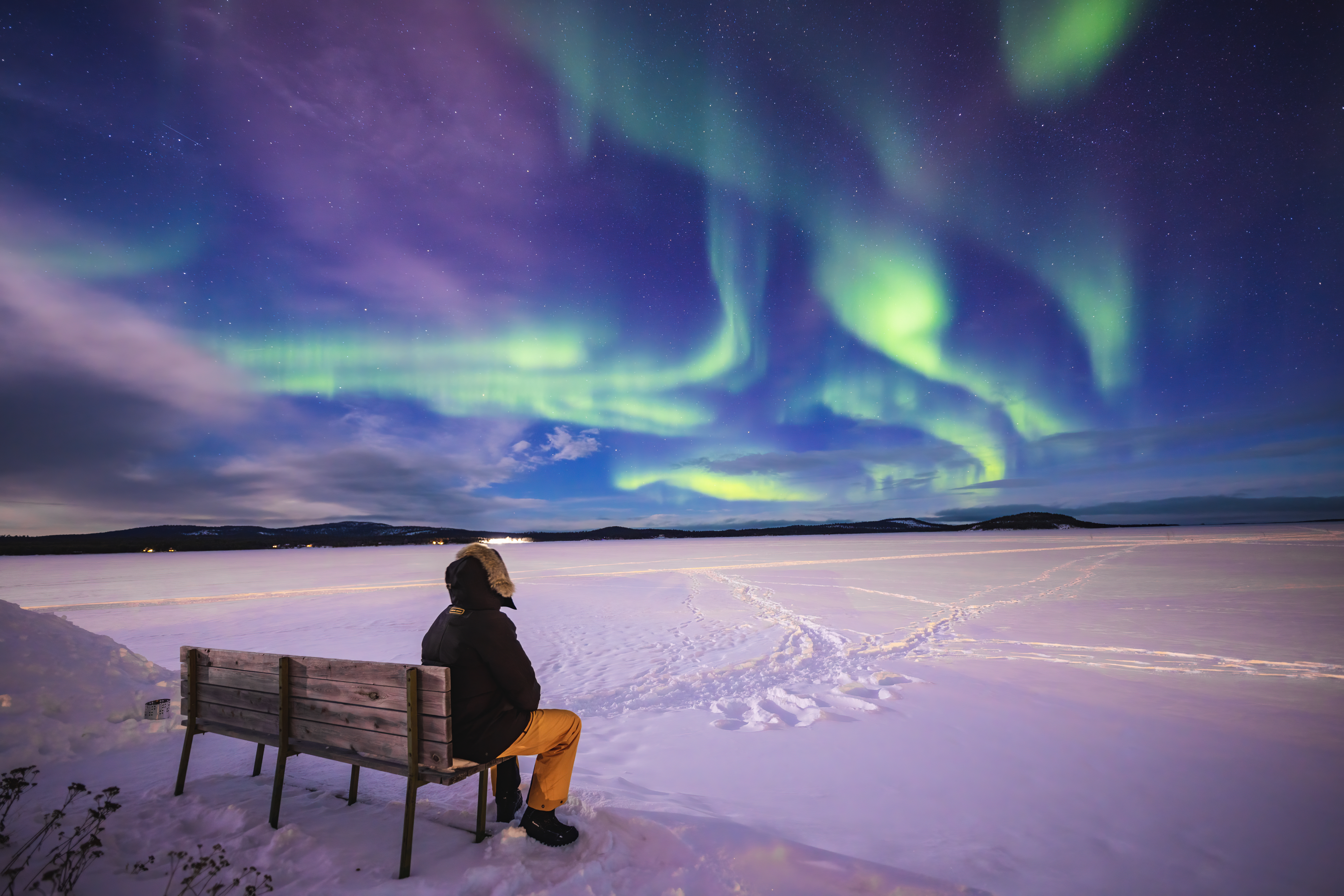 Person sitting on a bench, gazing at vibrant northern lights over a snowy landscape.
