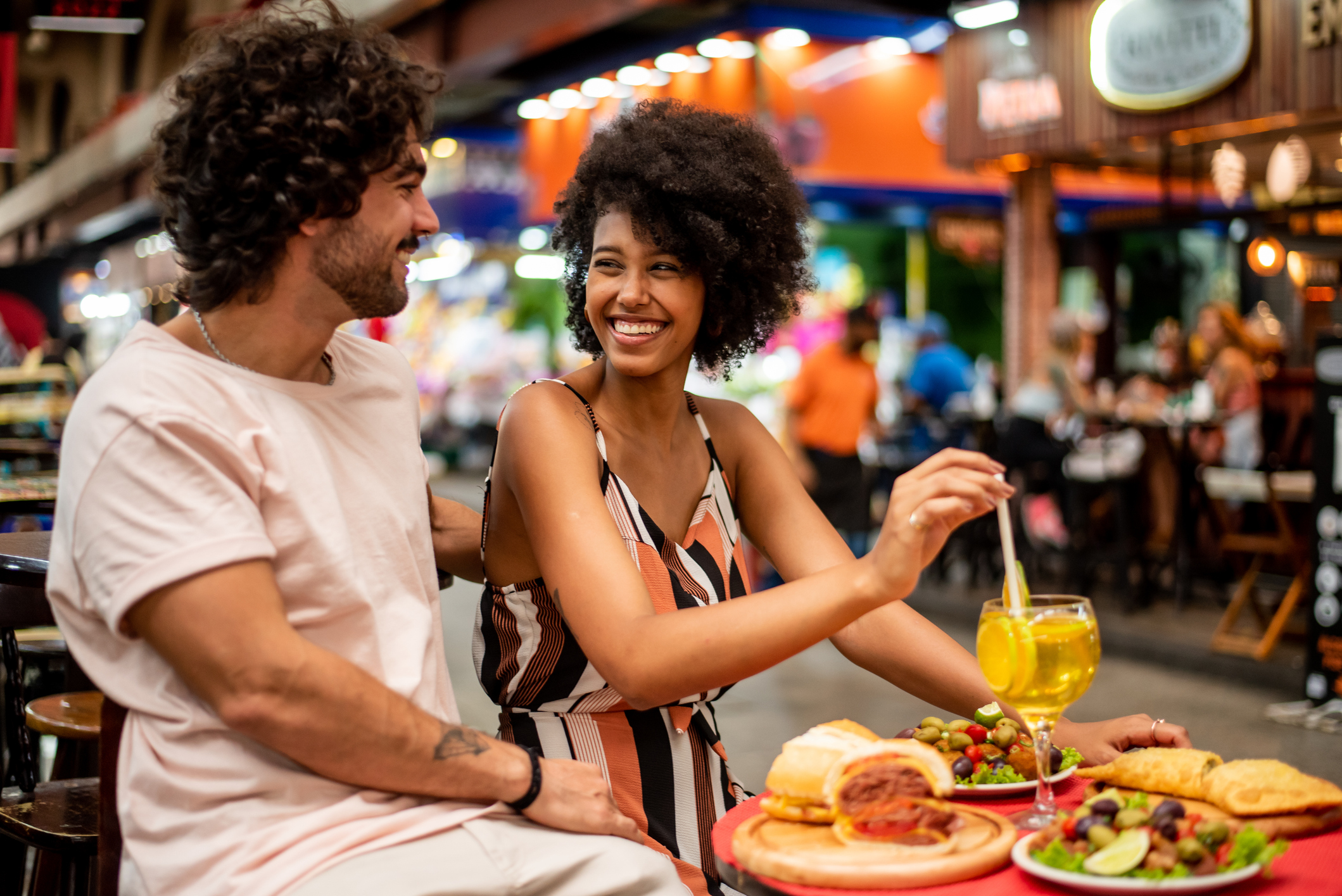 A couple enjoys a meal at a lively outdoor restaurant, smiling and sharing food and drinks in a vibrant atmosphere.