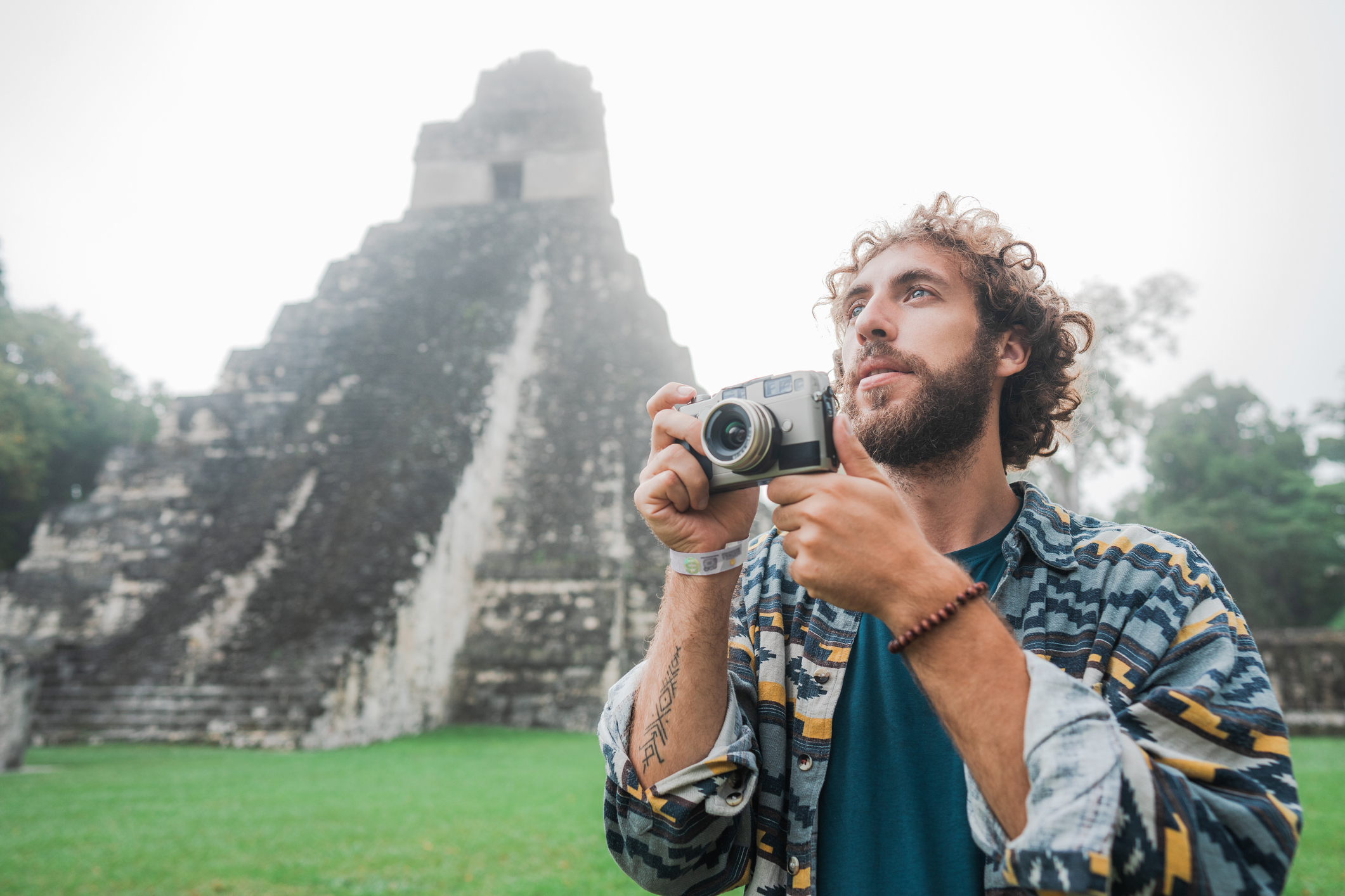 A man with curly hair holds a camera, standing in front of a Mayan pyramid in a misty landscape.