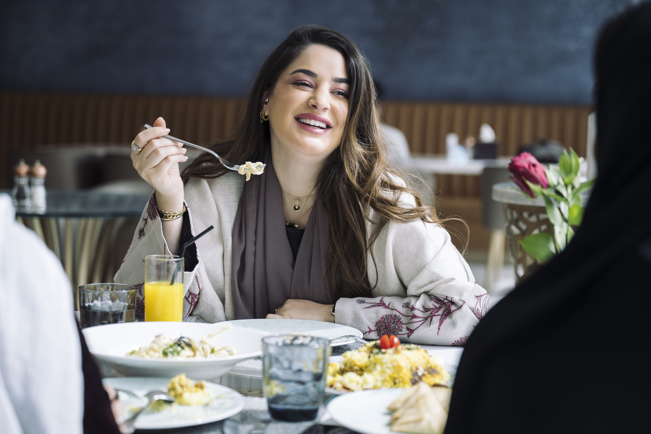 A woman smiles and enjoys a meal at a restaurant, surrounded by plates of food and drinks on the table.