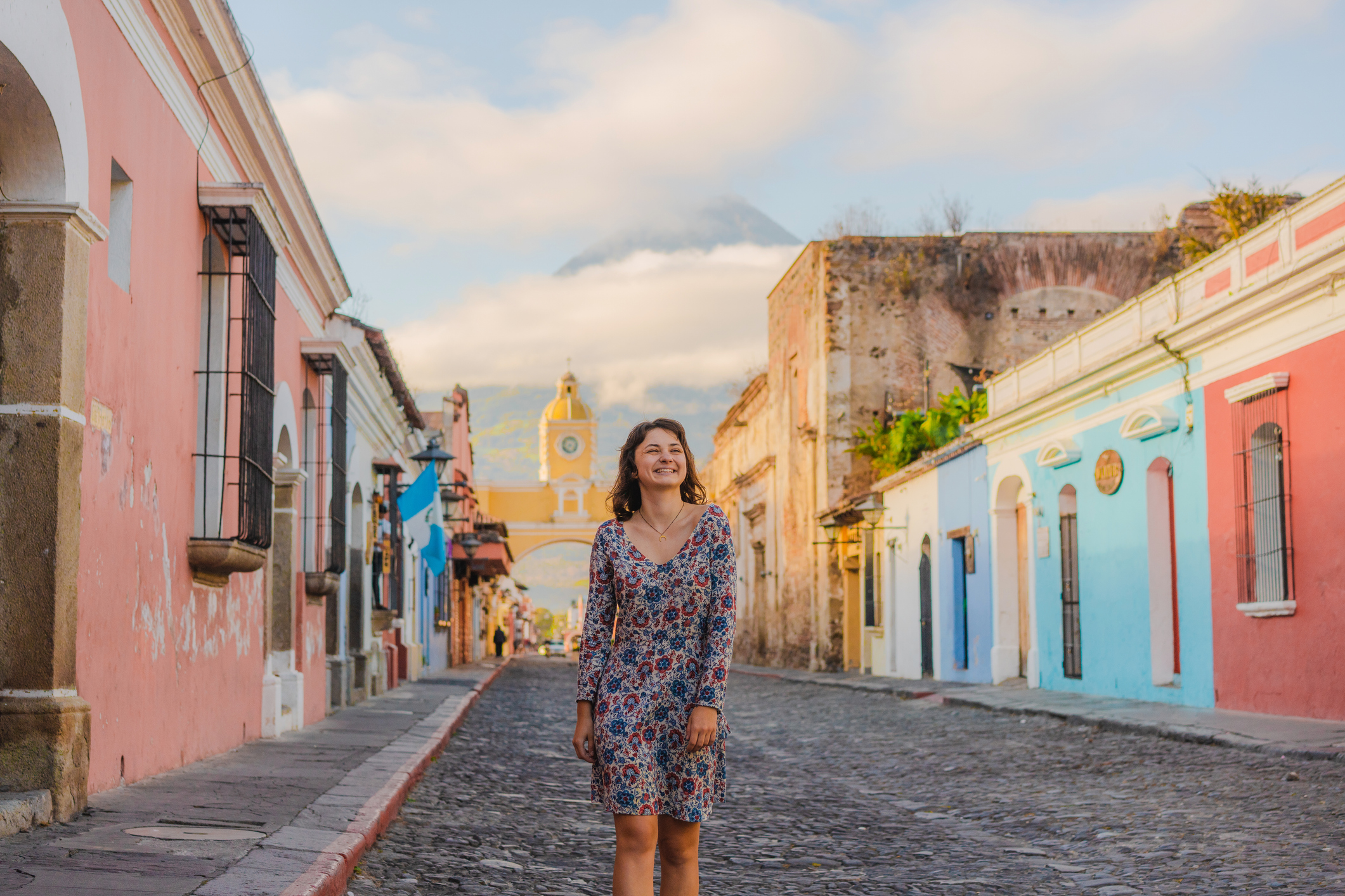 A woman stands on a cobblestone street lined with colorful buildings, with a mountain in the background under a clear sky.