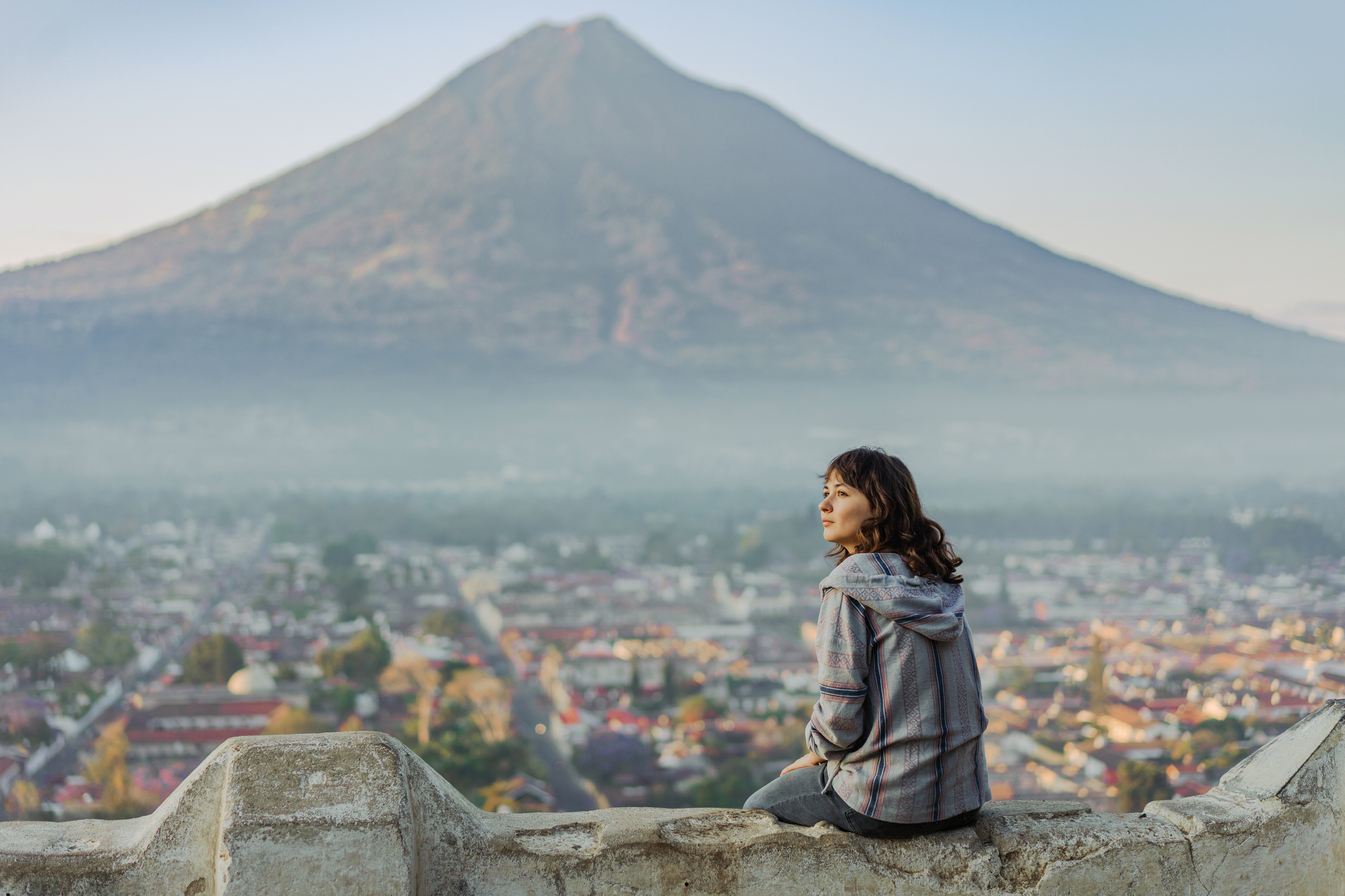 A person sits on a stone wall, overlooking a valley with a volcano in the background, surrounded by misty mountains.