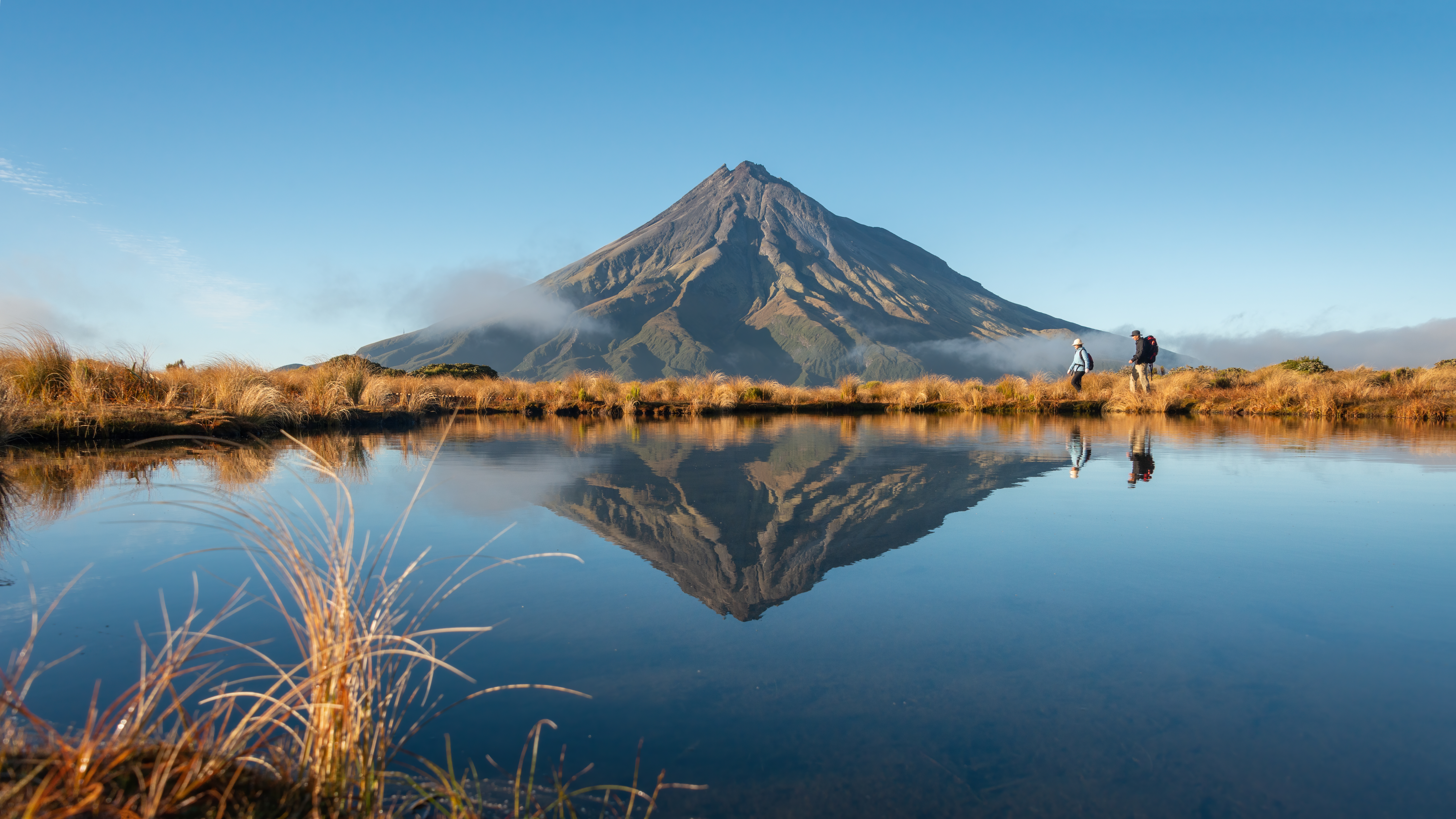 Two people hiking with the mountain reflected in the tarn in early morning sunshine