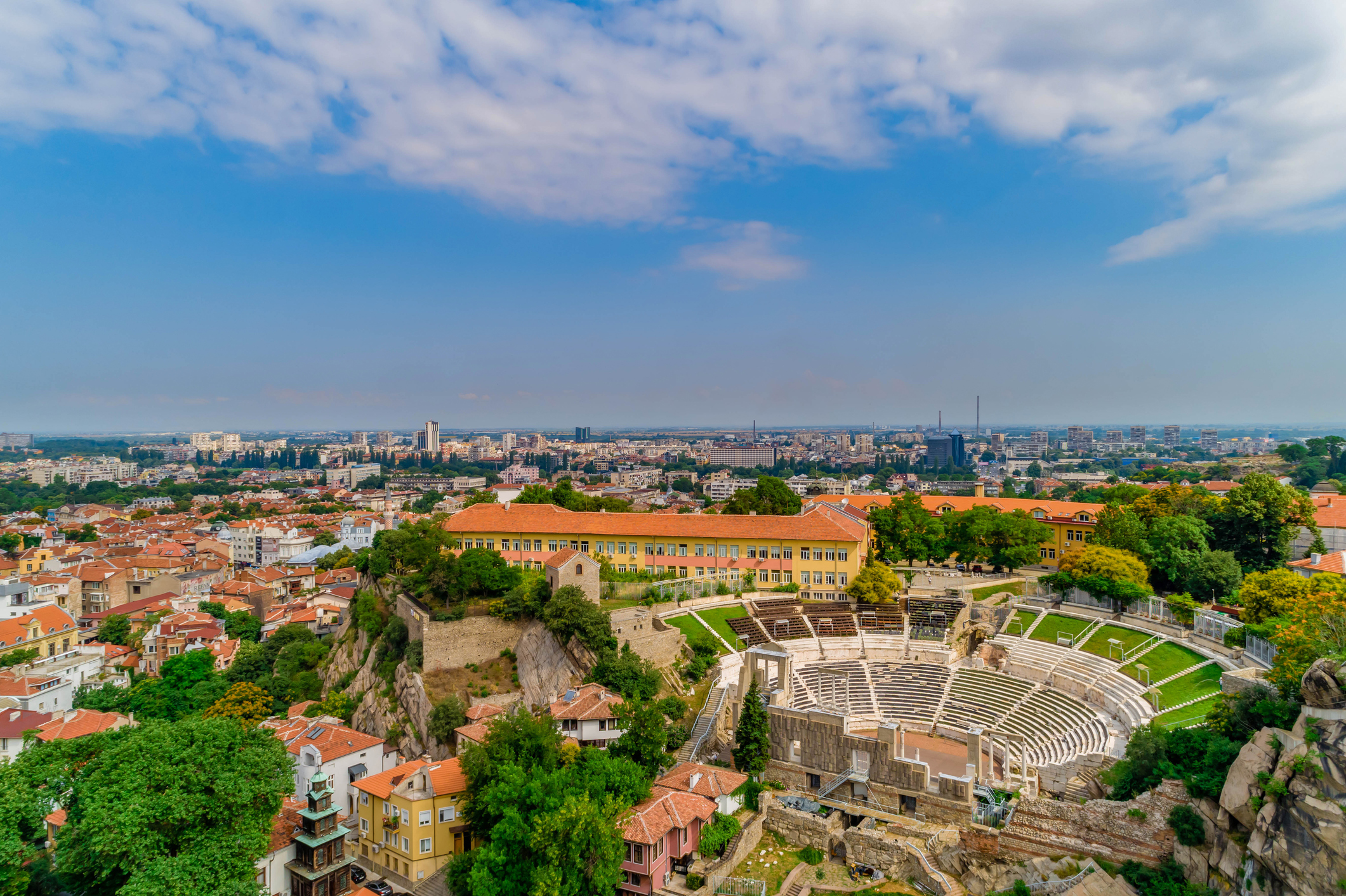 Aerial view of an ancient amphitheater surrounded by cityscape and greenery under a partly cloudy sky.