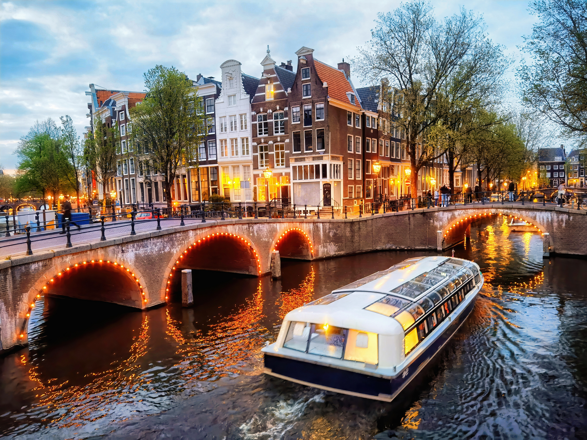A scenic view of a canal in Amsterdam with a boat and historic buildings lit up at dusk.