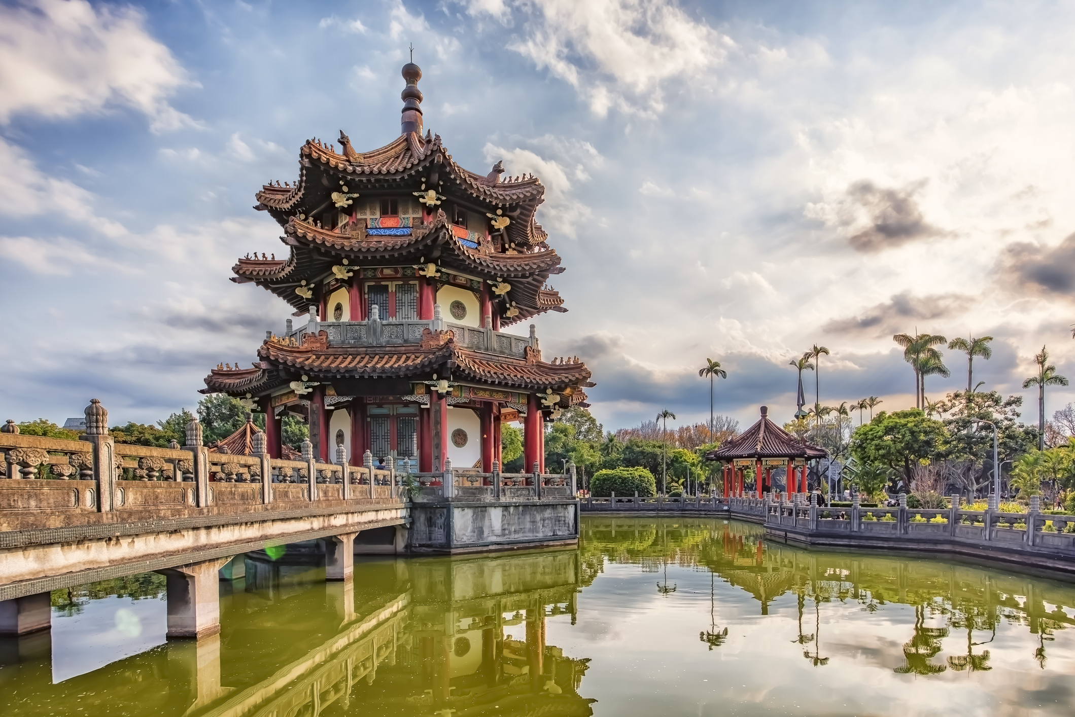 A traditional pagoda beside a tranquil pond, surrounded by lush greenery and a cloudy sky.