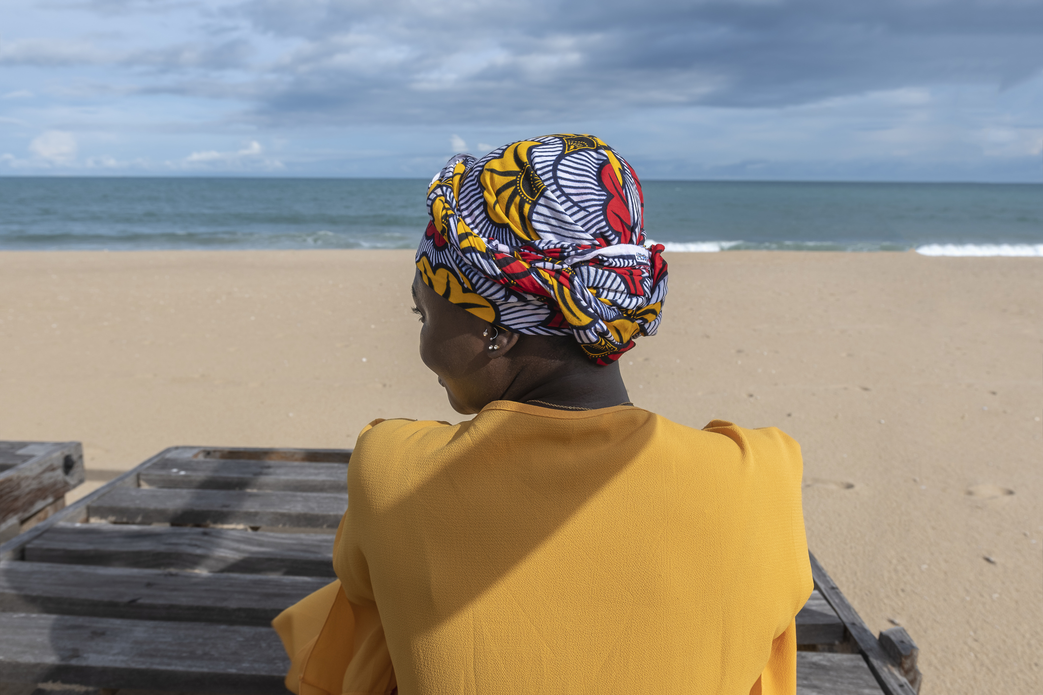A woman wearing a colorful headwrap sits by the beach, facing the ocean, with a sandy shore in the background.